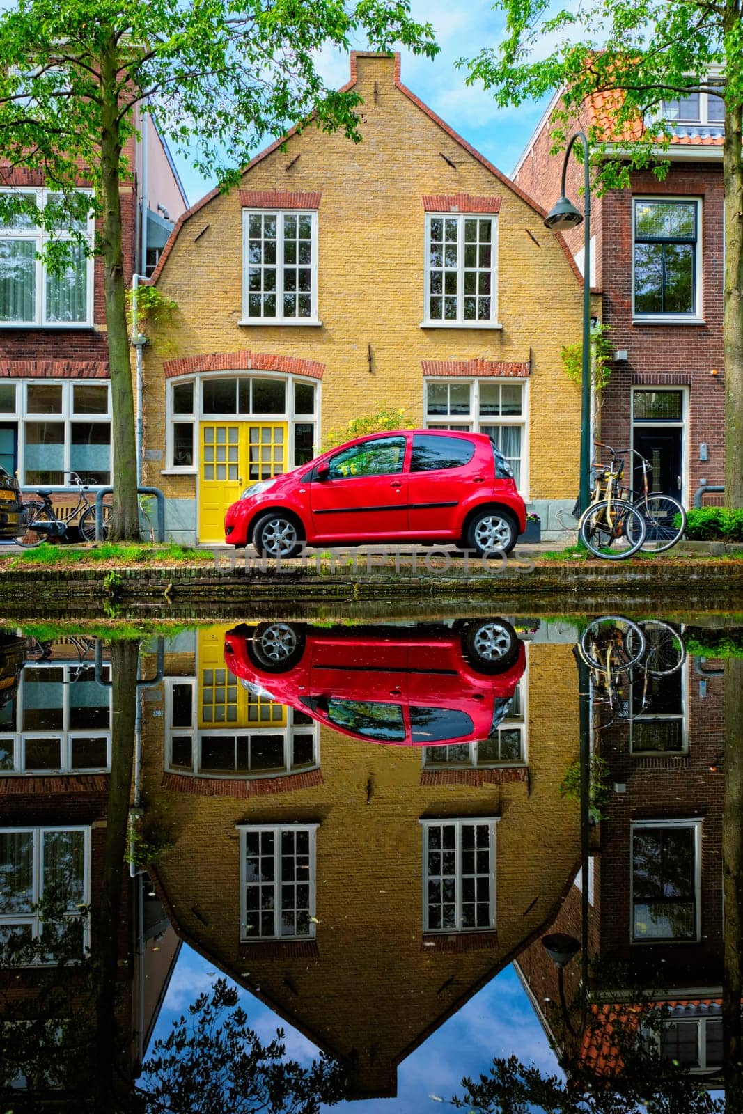 Red car on canal embankment in street of Delft with reflection and bicycle. Delft, Netherlands