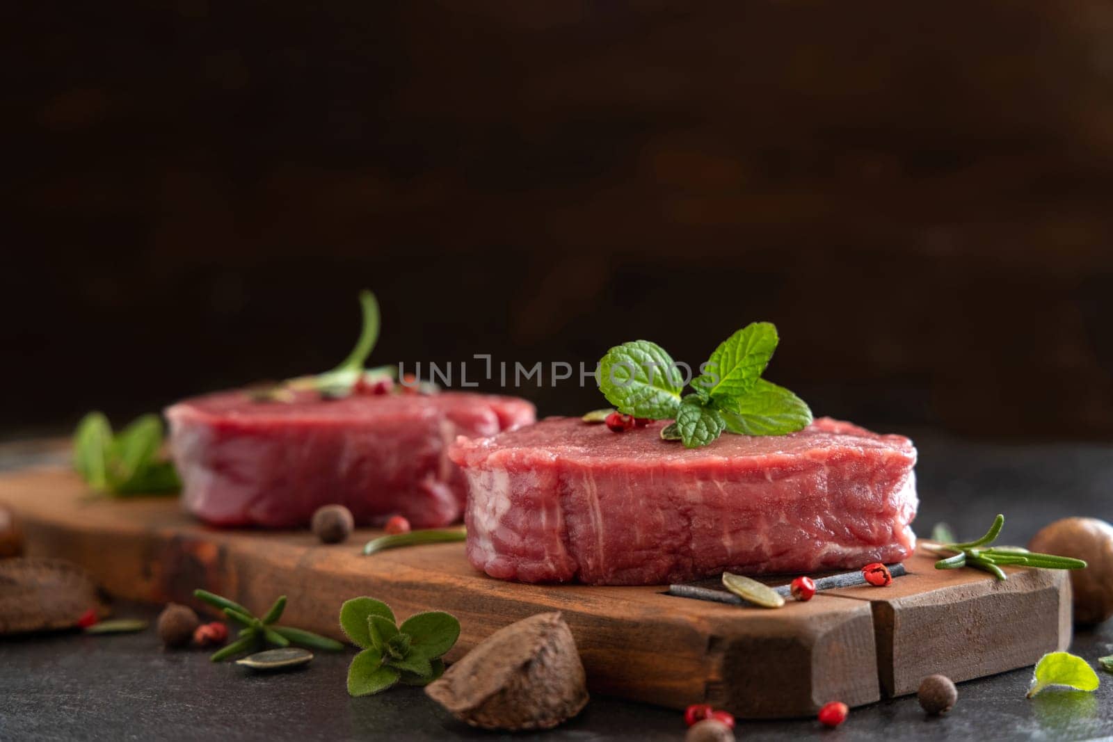 Raw beef fillet steaks on wooden board on dark table close up with copy space. Portioned filet mignons with spices and herbs. Concept of summer food, meat eaters.