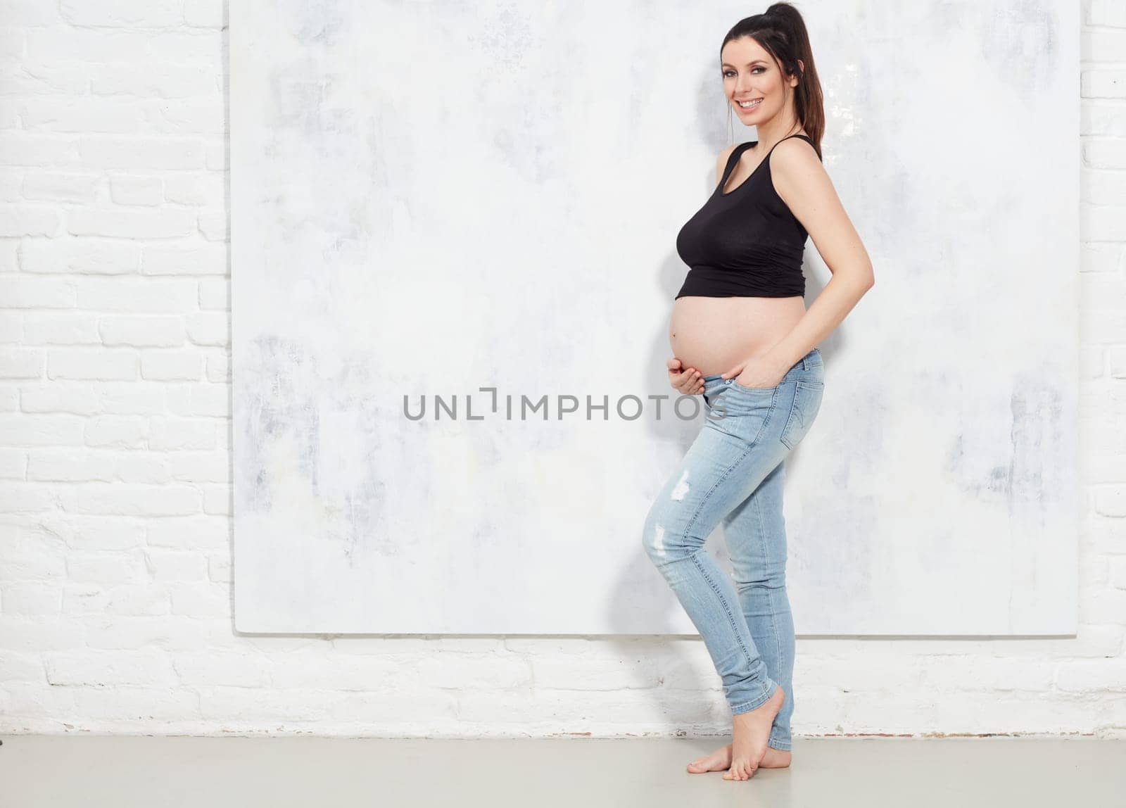Fashion portrait of happy pregnant woman with naked belly against white wall with copyspace.