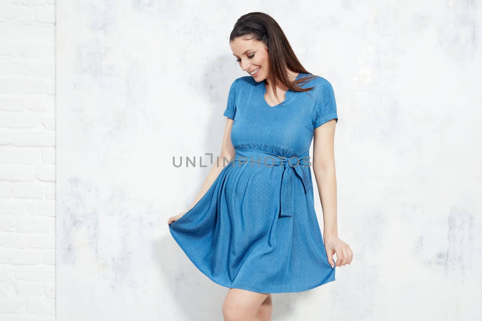 Fashion portrait of happy pregnant woman in blue summer dress against white wall with copyspace.