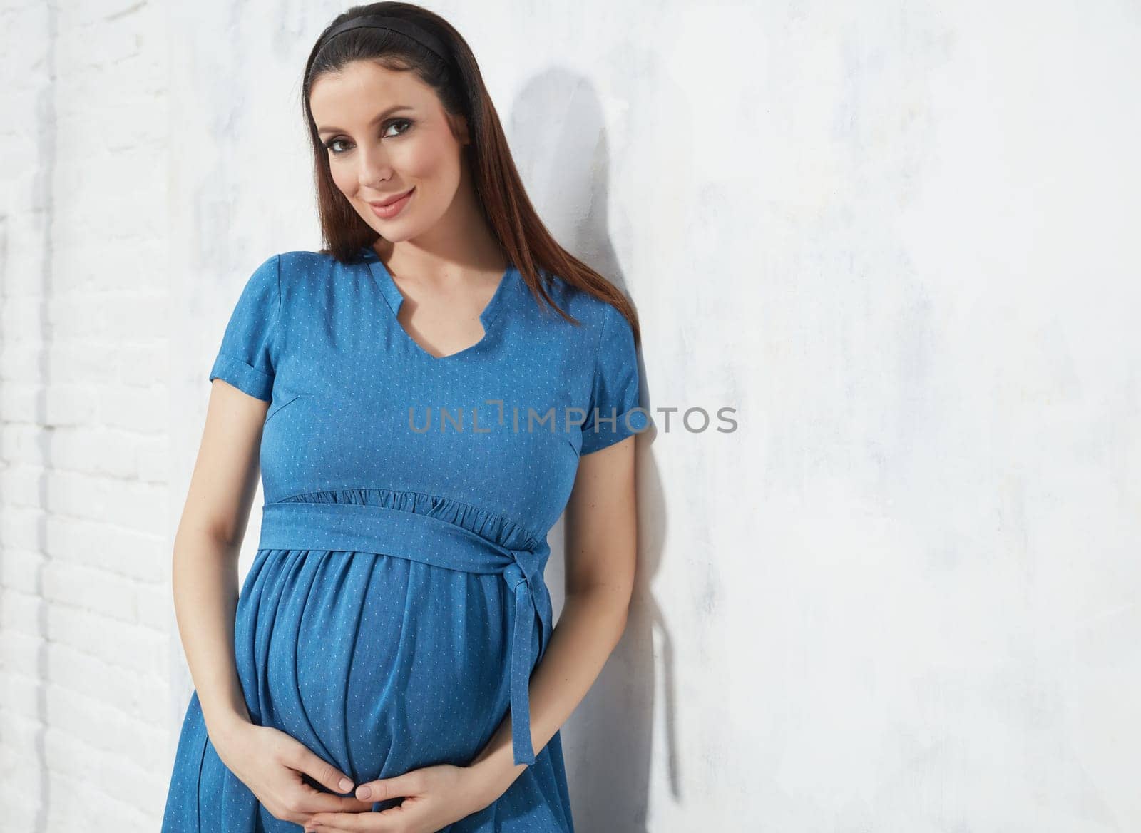 Fashion portrait of happy pregnant woman in blue summer dress against white wall with copyspace.