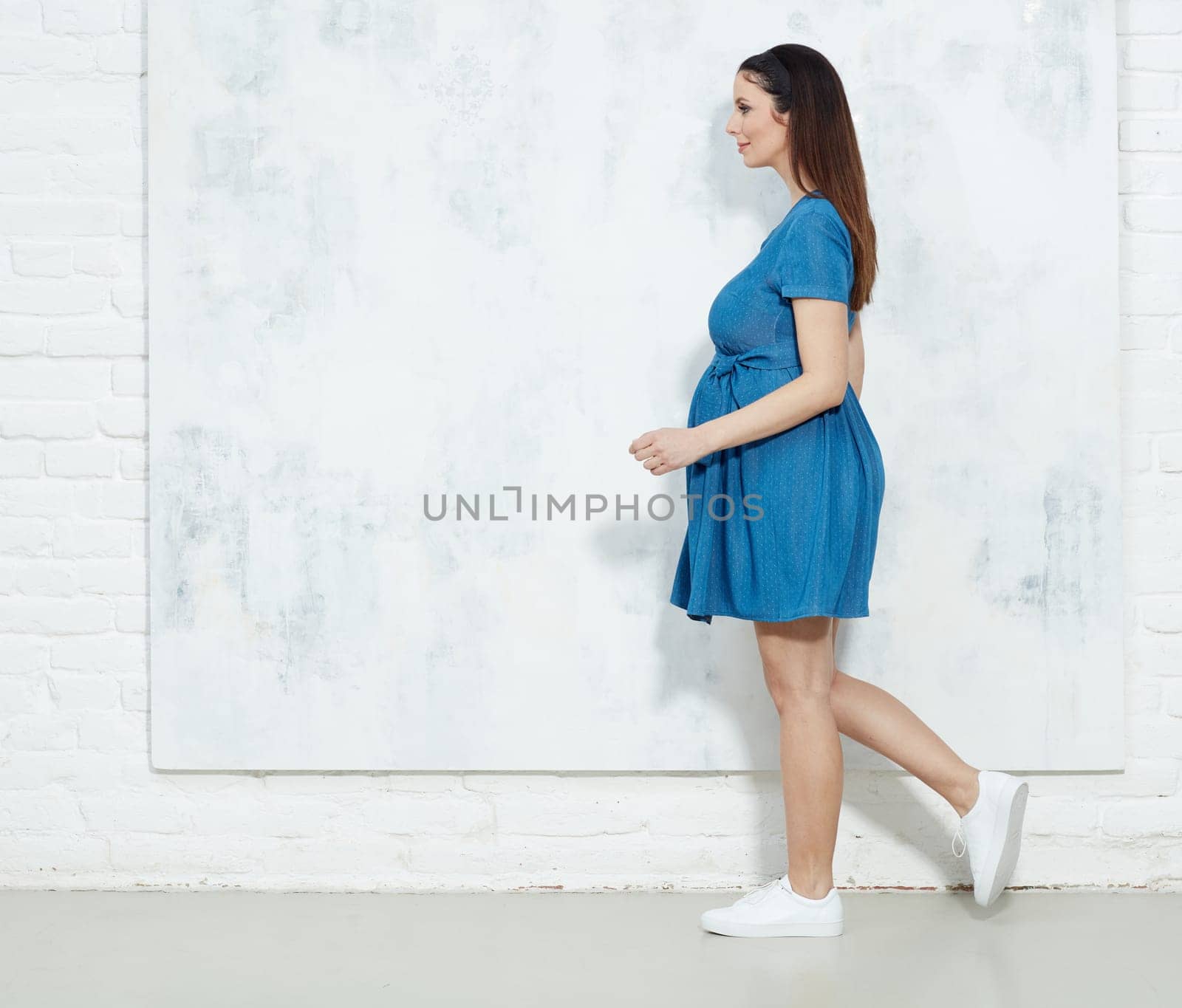 Fashion portrait of happy pregnant woman in blue summer dress against white wall with copyspace.