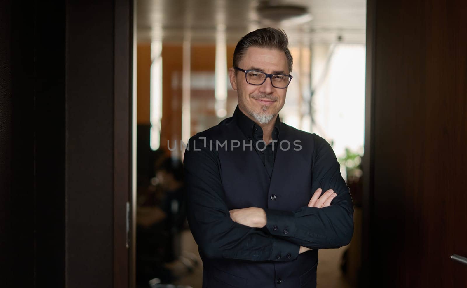 Business portrait - businessman standing in office, smiling. Happy middle aged, mid adult man in black jacket.
