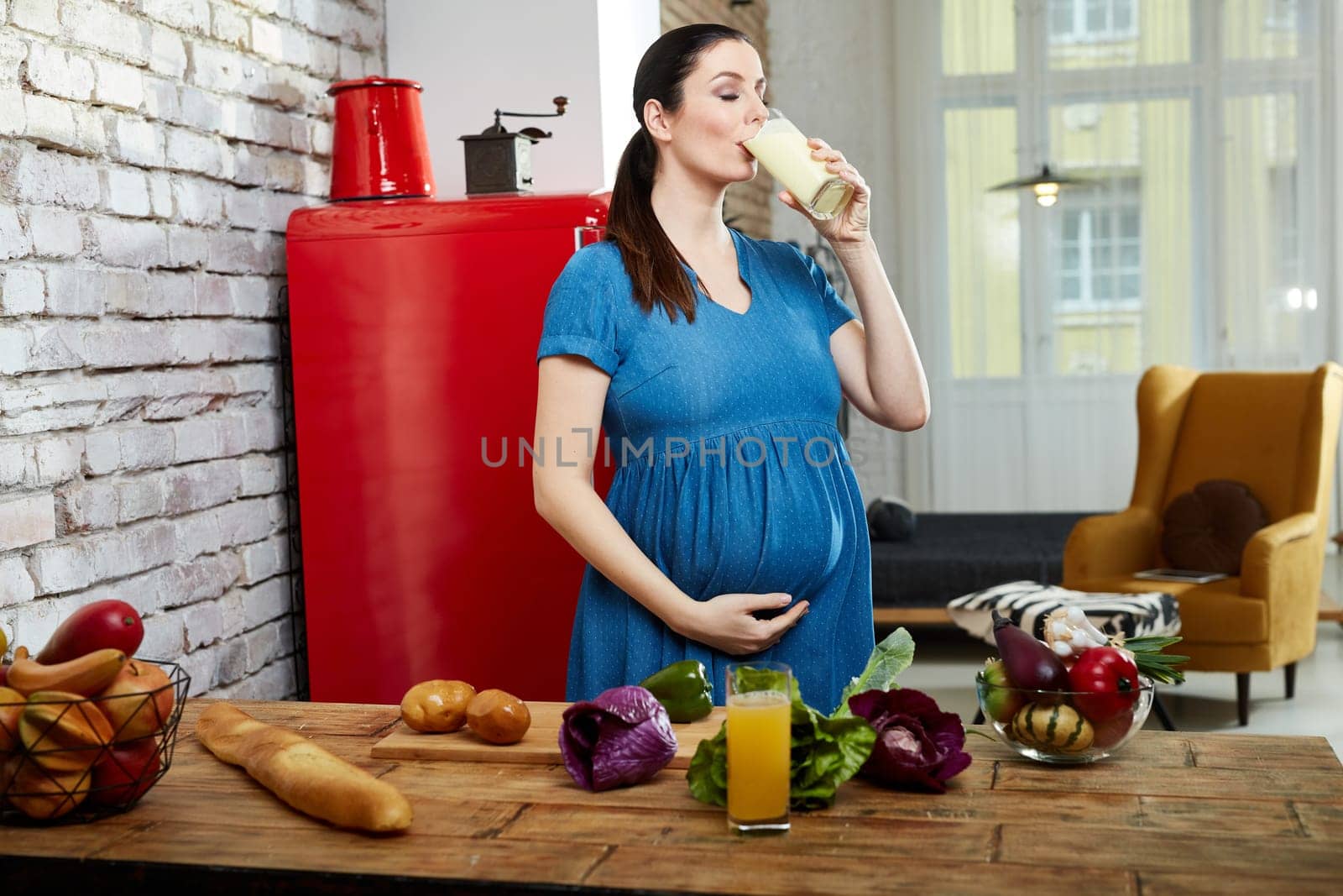 Happy pregnant woman at home in kitchen with living room in background drinking shake made of healthy vegetables.