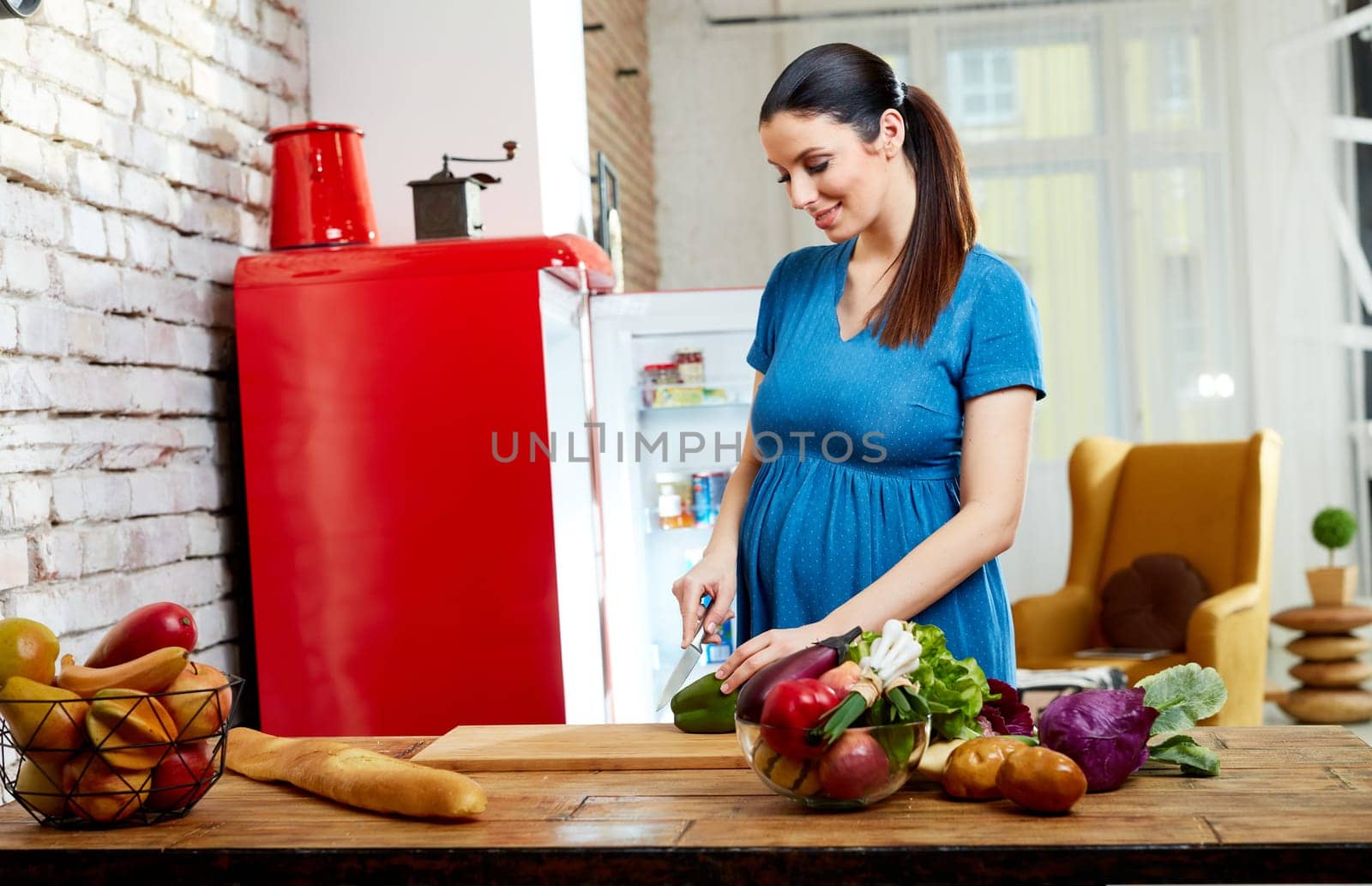 Happy pregnant woman at home in kitchen with living room in background preparing salad, eating healthy food from vegetables.