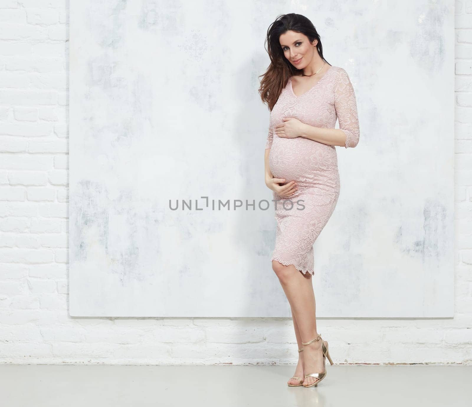 Fashion portrait of happy pregnant woman in elegant dress against white wall with copyspace.