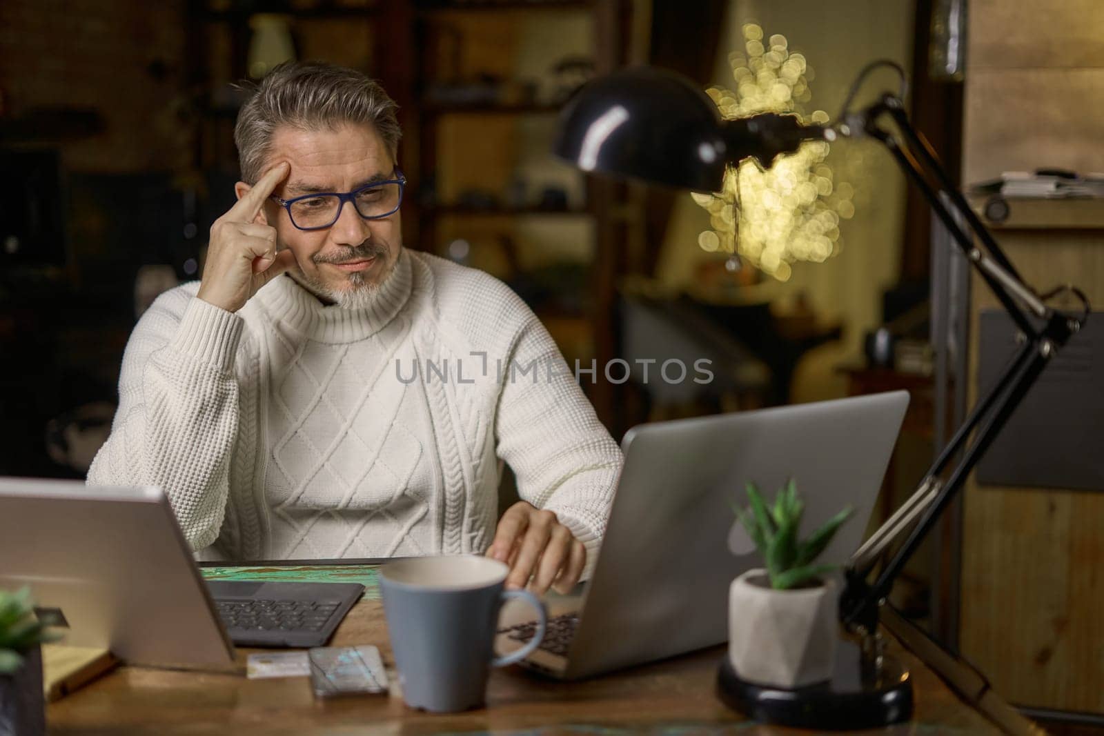 Happy mid adult man working with laptop computer in home office, glasses, gray hair. Winter, Christmas, dark cosy room.
