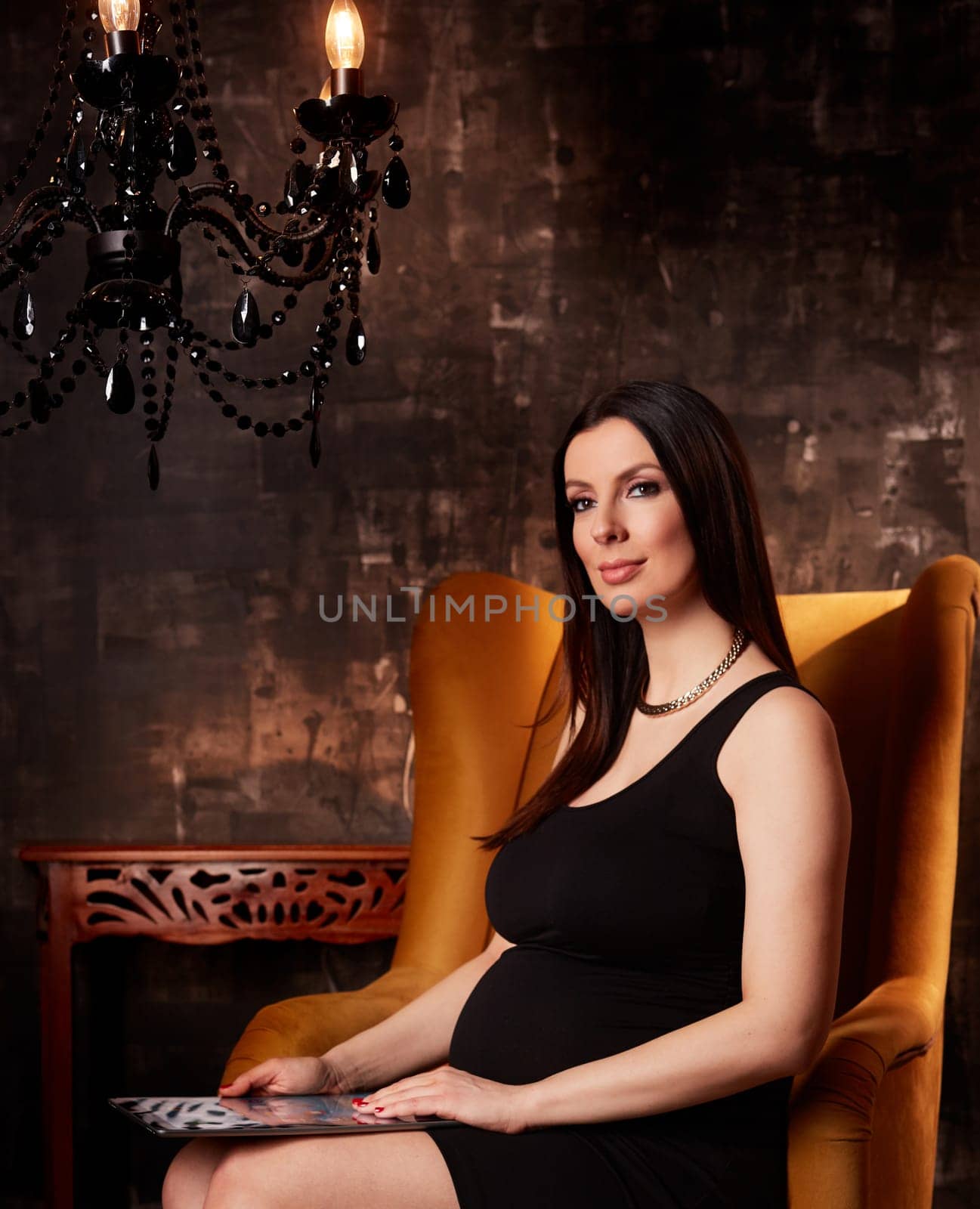 Beautiful pregnant woman in elegant black dress sitting in hotel lobby holding handheld tablet.