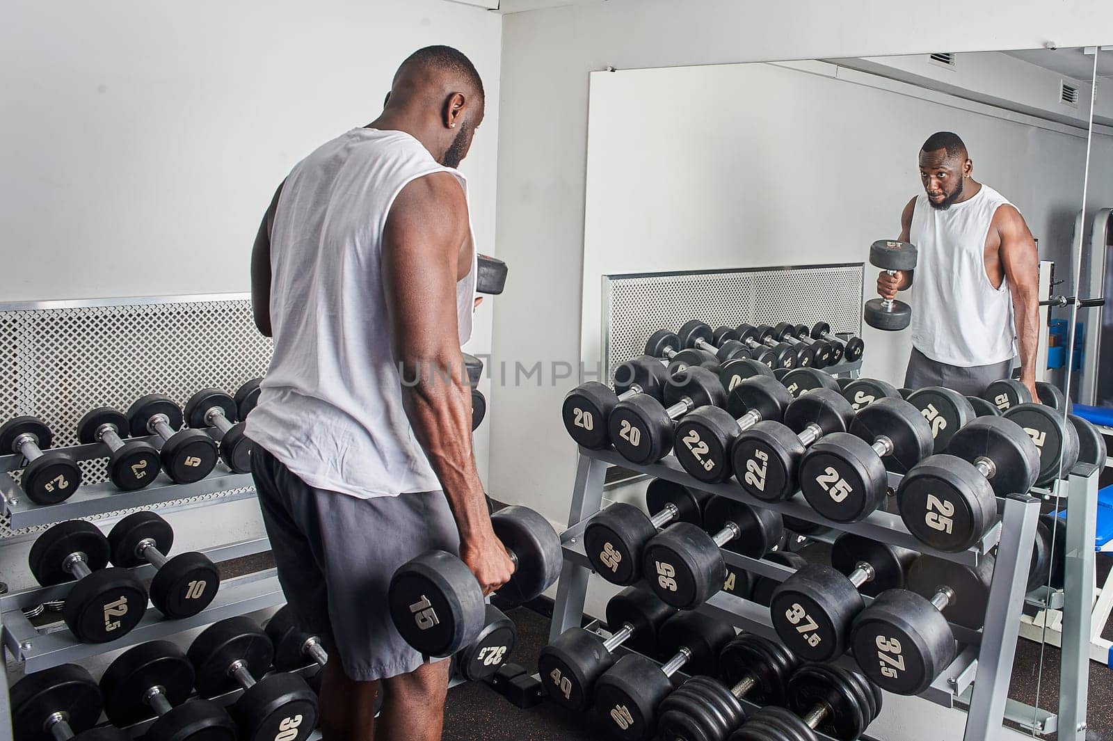 An African American man shakes his biceps with dumbbells while standing in front of a mirror. High quality photo