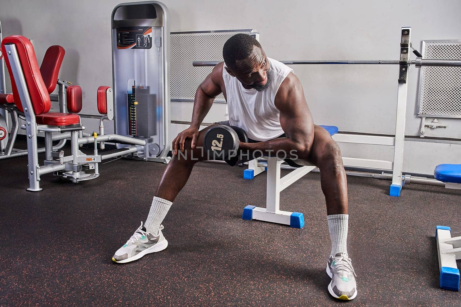 An African American man shakes his biceps while sitting on a bench in the gym. High quality photo