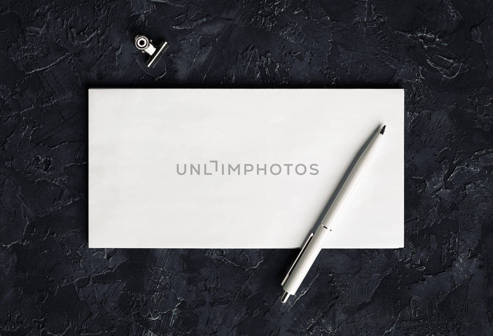 Blank envelope, pen and clip on black plaster background. Branding mock up. Top view.