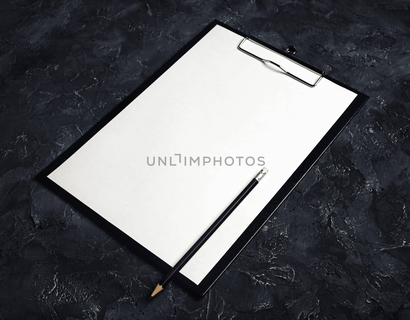 Photo of clipboard with a blank sheet of paper and pencil on black plaster background. Mock-up for branding identity.