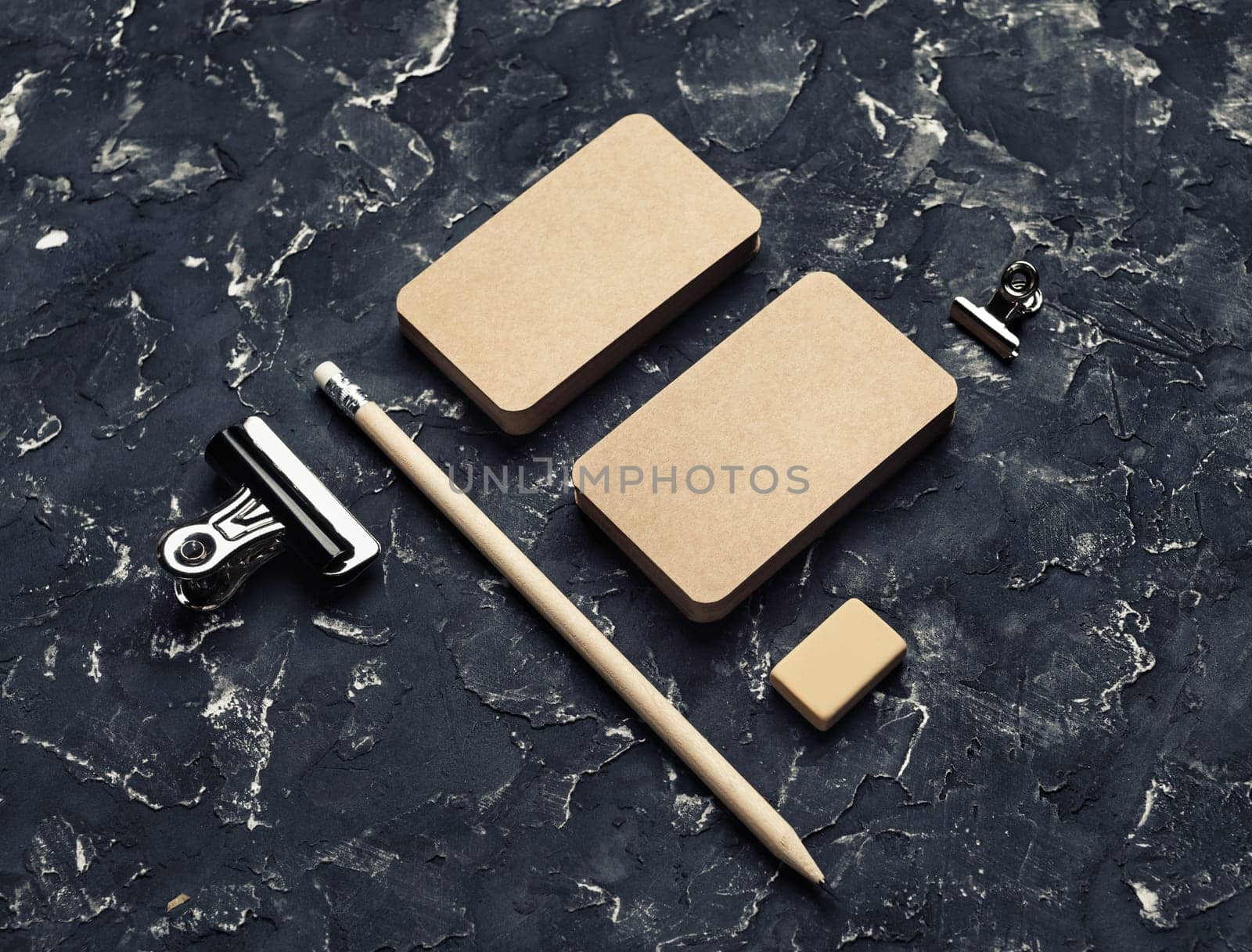 Blank stationery template. Blank kraft business cards, pencil, eraser and metal clips on black plaster background.