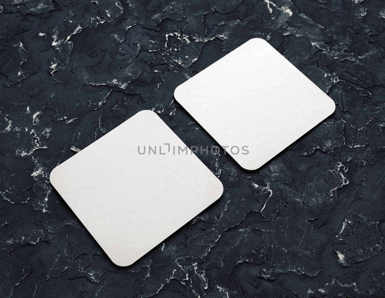 Two blank square beer coasters on black plaster background.