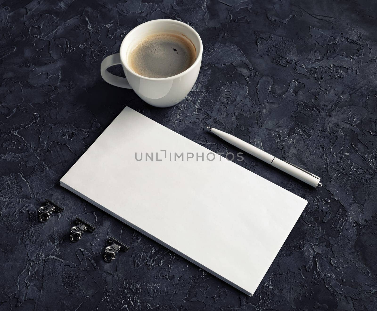 Blank white paper envelope, coffee cup, pen and metal clips on black plaster background.