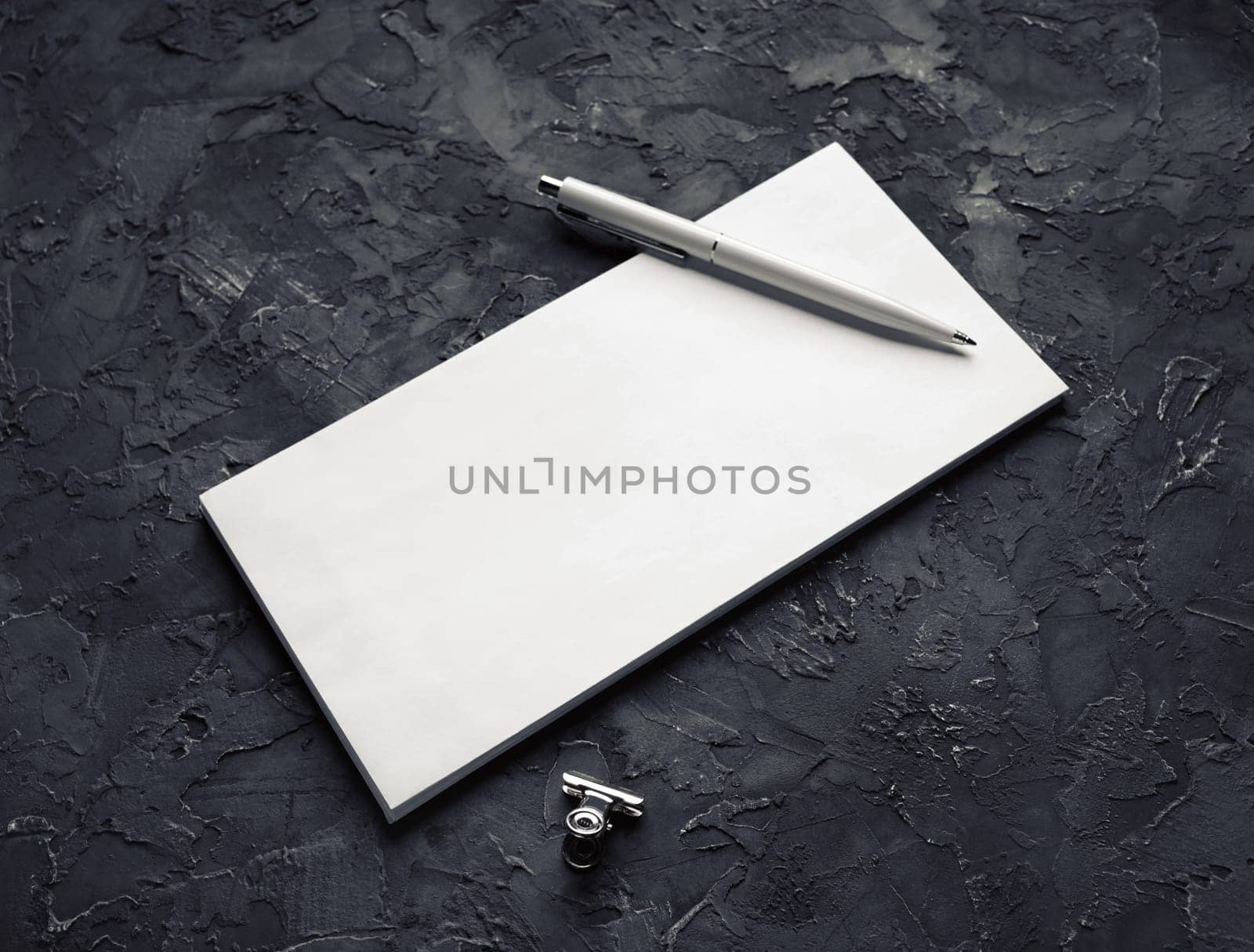 Photo of blank paper envelope, pen and metal clip on black plaster background.