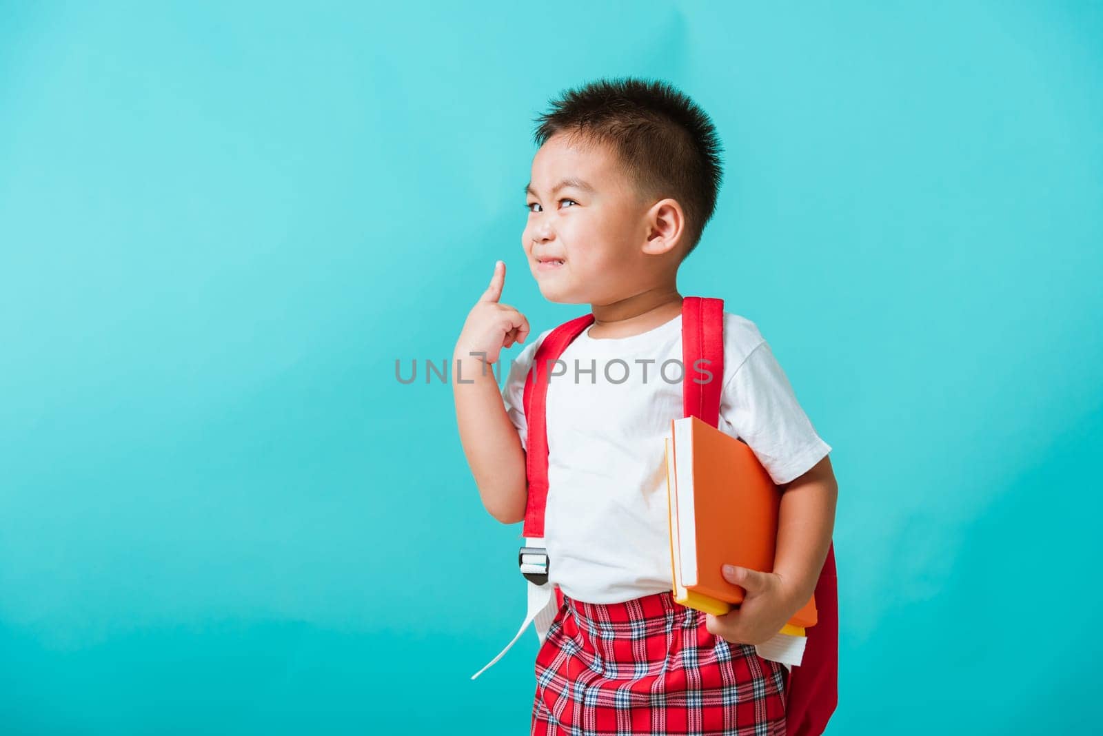 Back to school concept. Portrait Asian happy funny cute little child boy smile hug books and point finger to side away space, isolated blue background. Kid from preschool kindergarten with school bag