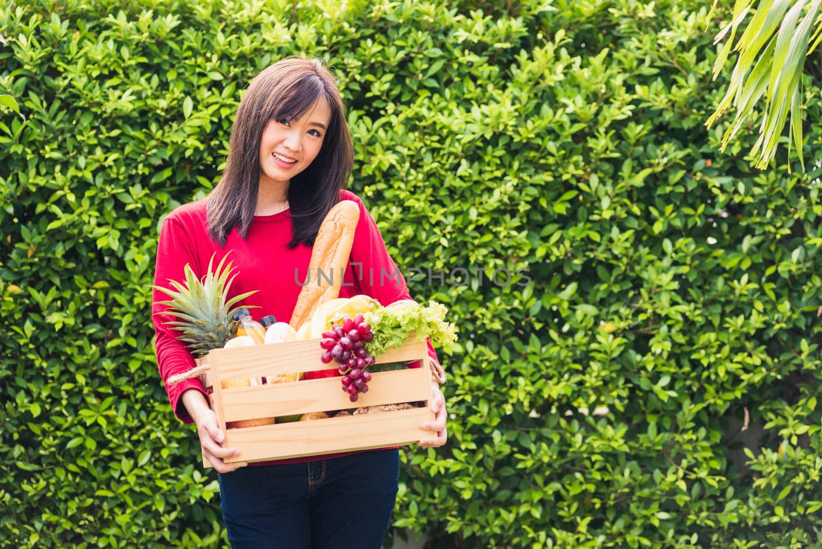 Portrait of Asian beautiful young woman farmer standing she smile and holding full fresh food raw vegetables fruit in a wood box in her hands on green leaves background