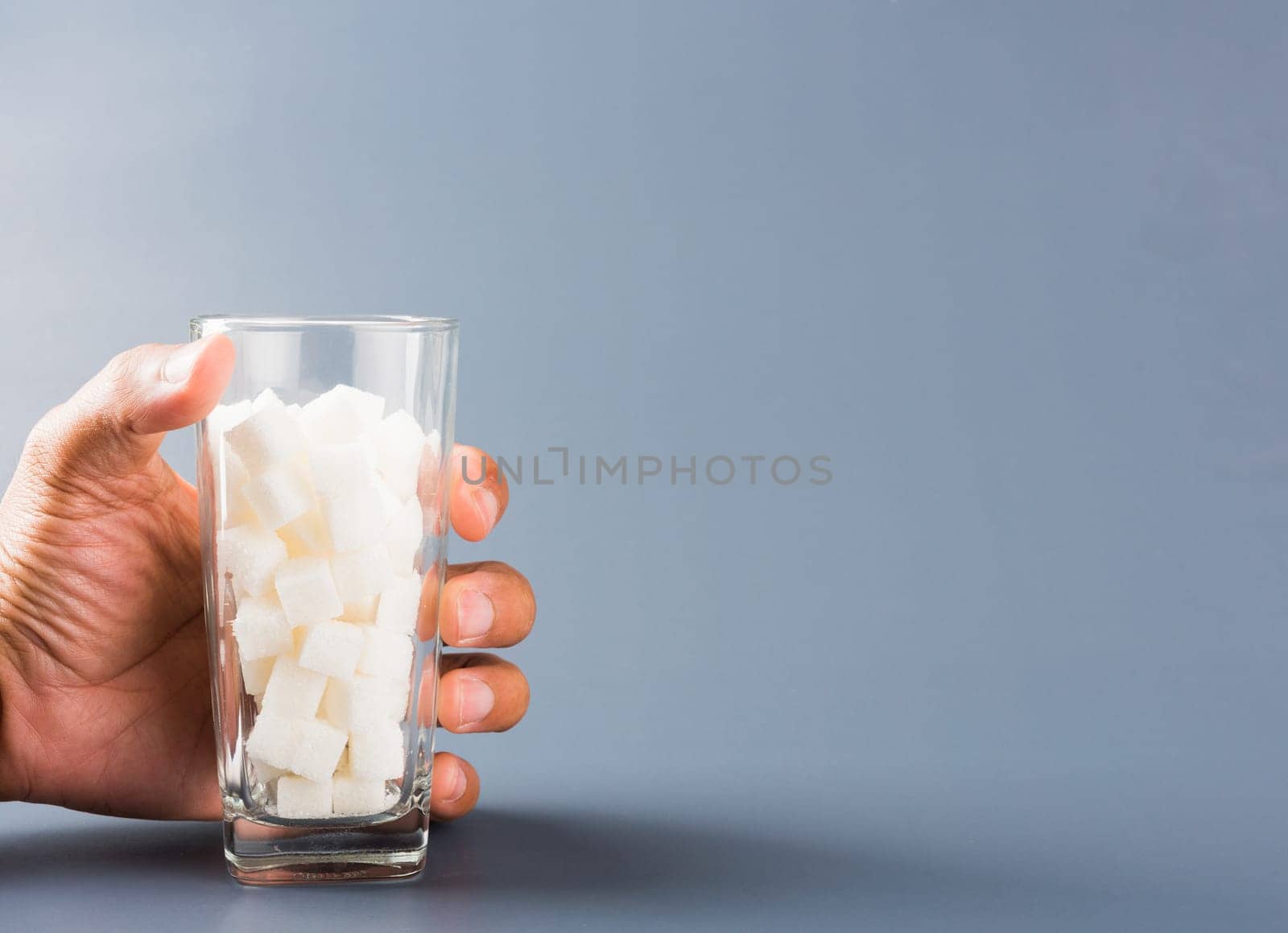 Hand hold on glass full of white sugar cube sweet food ingredient, isolated on gray background, health high blood risk of diabetes and calorie intake concept and unhealthy drink