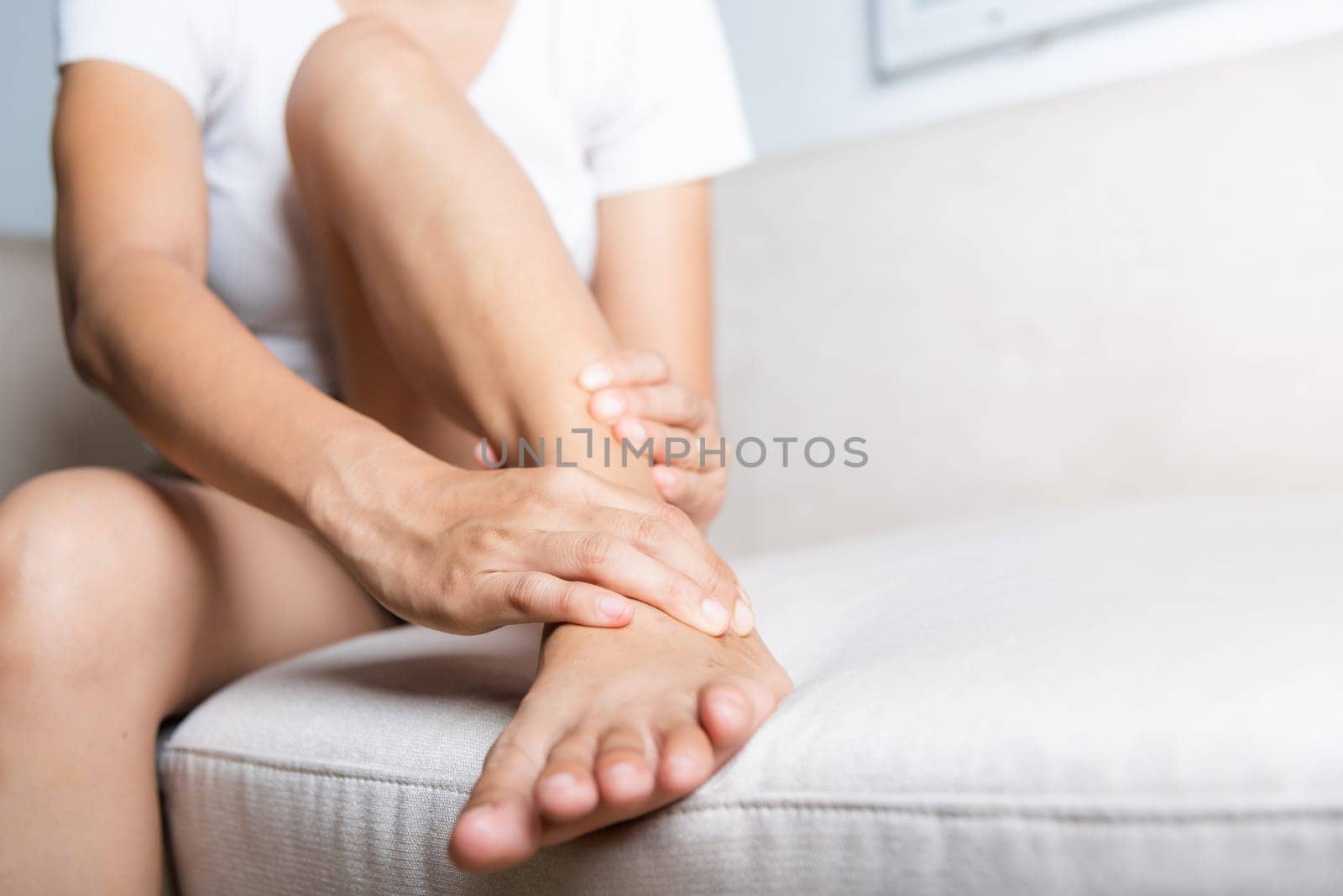 Foot pain, Asian woman sitting on sofa hold her ankle injury feeling pain in her foot at home, female suffering from feet ache use hand massage relax muscle from ankle interior, Healthcare and medical