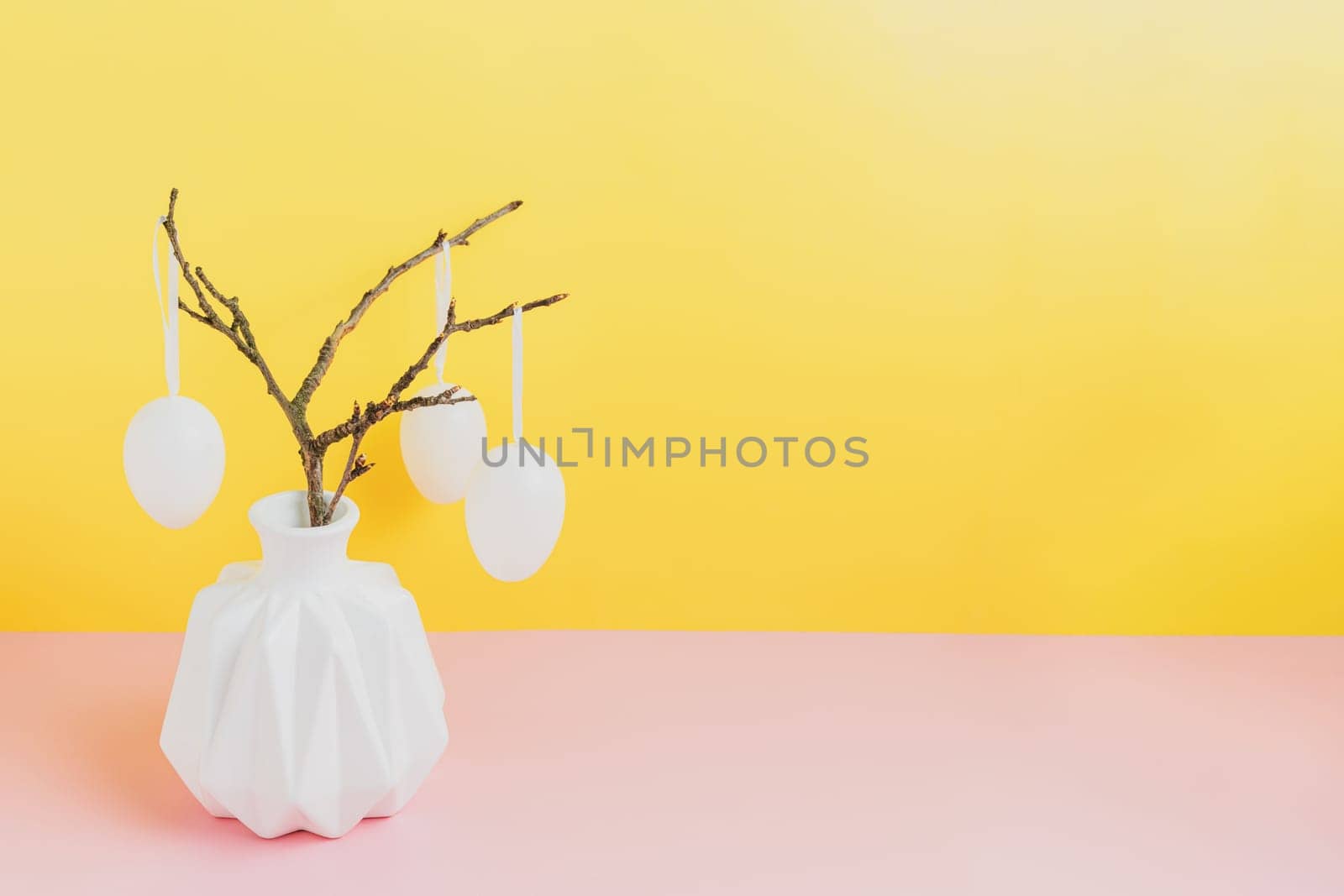Happy Easter holiday, coming spring concept. Tree branches, flower twigs in white vase or jar with hanging eggs. Yellow and pink table background with copy space for ad or text. Easter home decoration