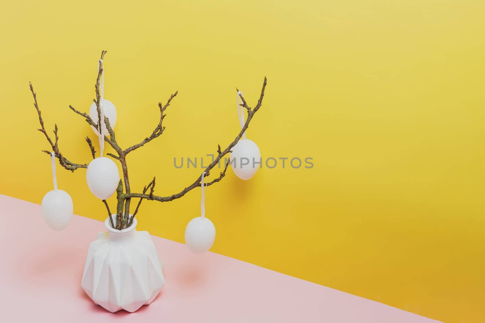 Happy Easter holiday, coming spring concept. Tree branches, flower twigs in white vase or jar with hanging eggs. Yellow and pink table background with copy space for ad or text. Easter home decoration