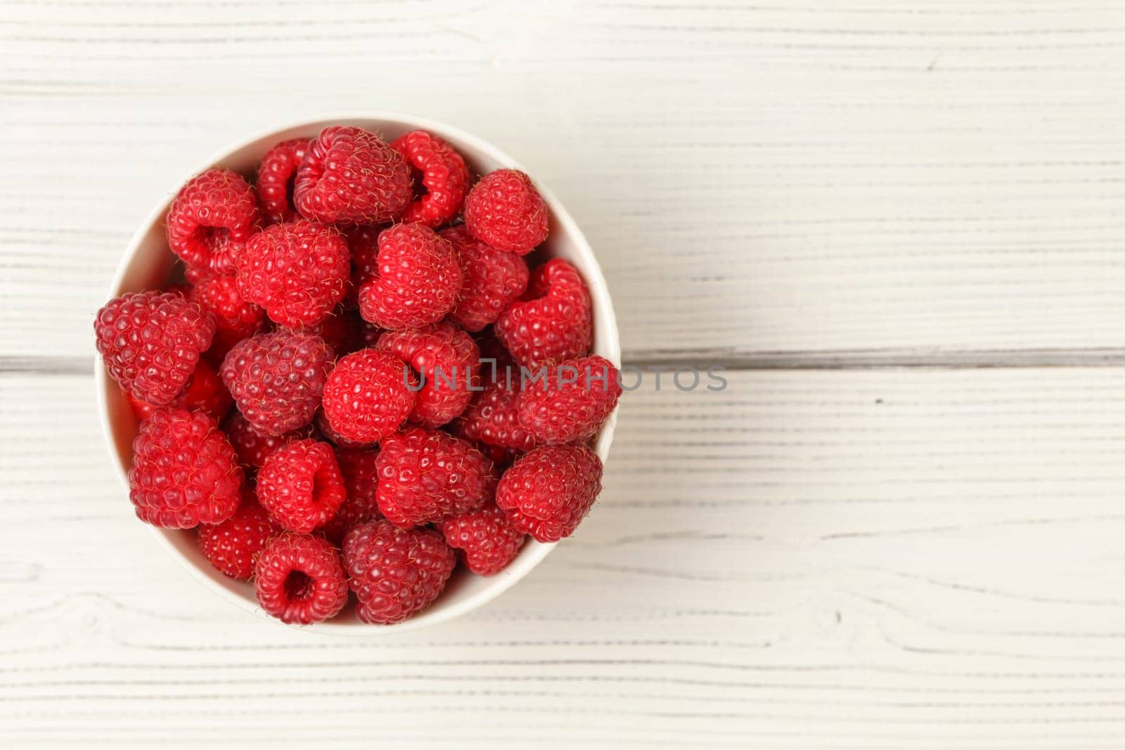 Tabletop view, small porcelain cup filled with raspberries on white boards desk.