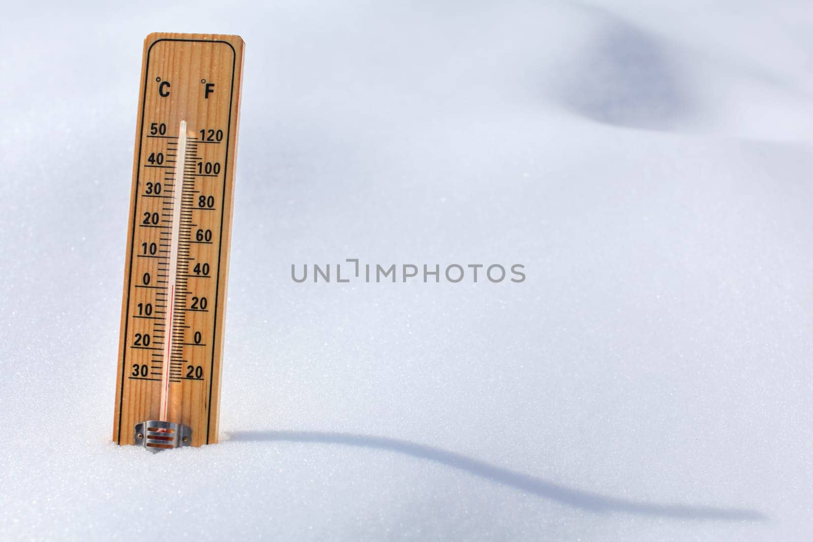 Wooden thermometer standing in the snow, sun casting curvy shadow, red column showing 0 degrees Celsius temperature. Winter / snow coming concept.