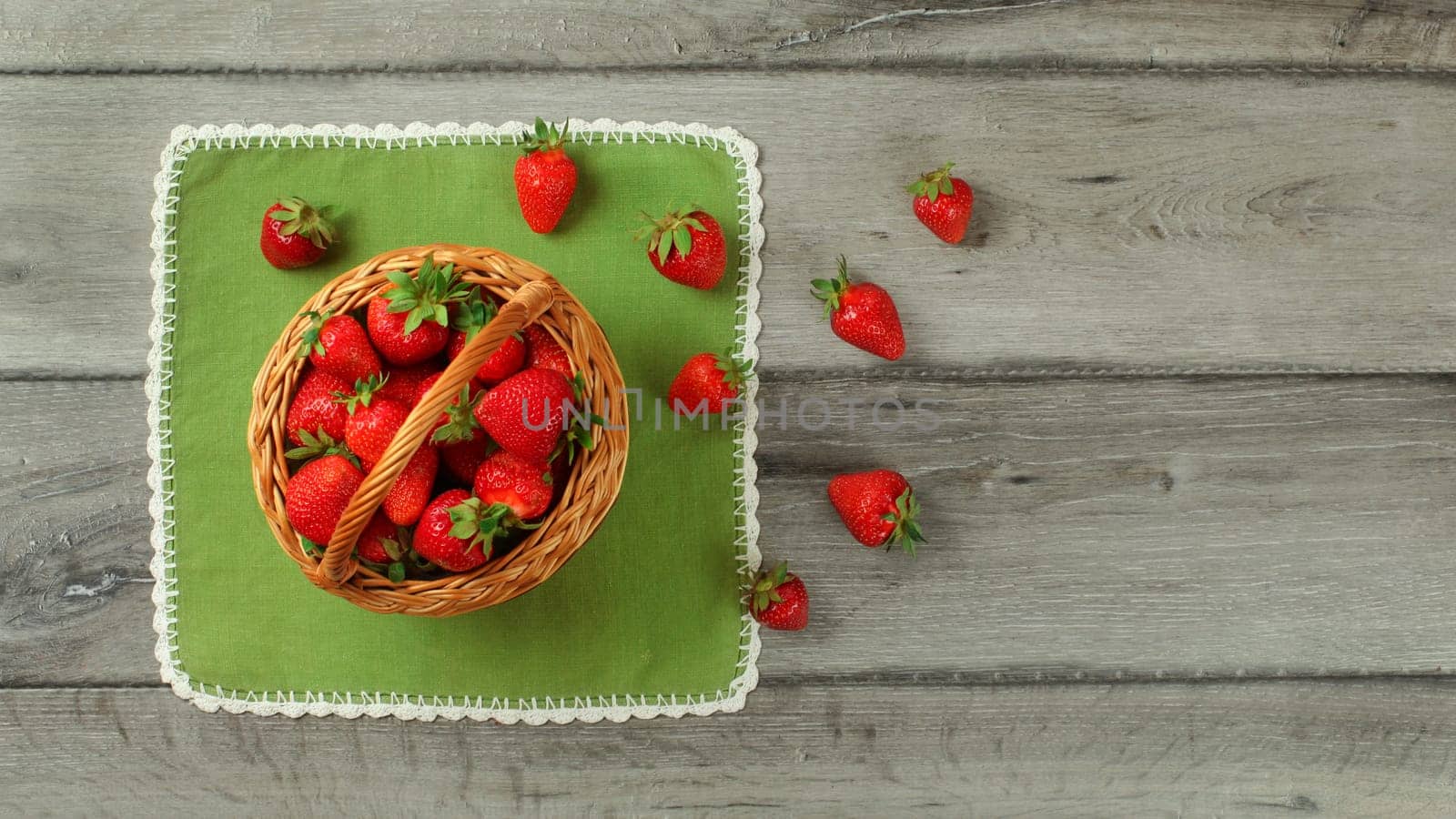 Tabletop view, basket with strawberries, some of them spilled on green tablecloth under it.