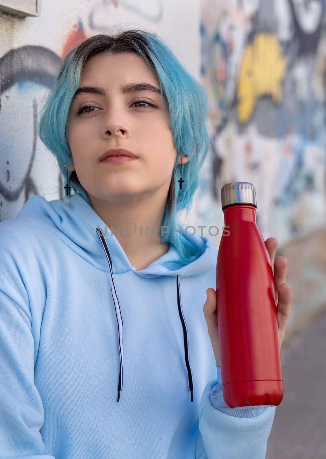 Teenager in blue hoodie with red water bottle looking aside. Blue haired teen girl staying outdoors near graffiti wall. Clothing and reusable bottle mockup. Hipster and adolescence concept