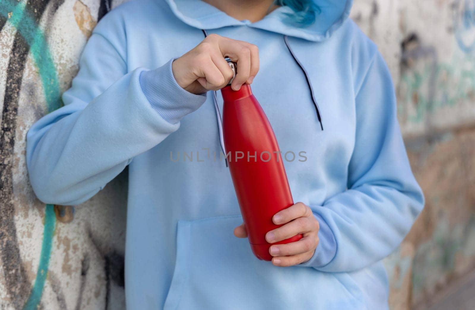 Young woman in blue hoodie with red water bottle staying outdoors near graffiti wall. Clothing and reusable bottle mockup. Hipster and adolescence concept