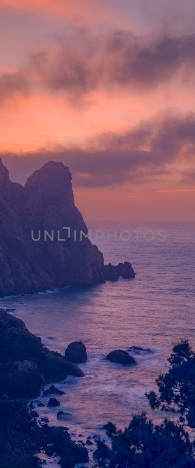 Beautiful seascape at sunset with clouds in the sky. In the foreground are dark branches of trees, the sea reflects the lilac sunset sky, the mountain is also in a sunset haze of heavenly color.