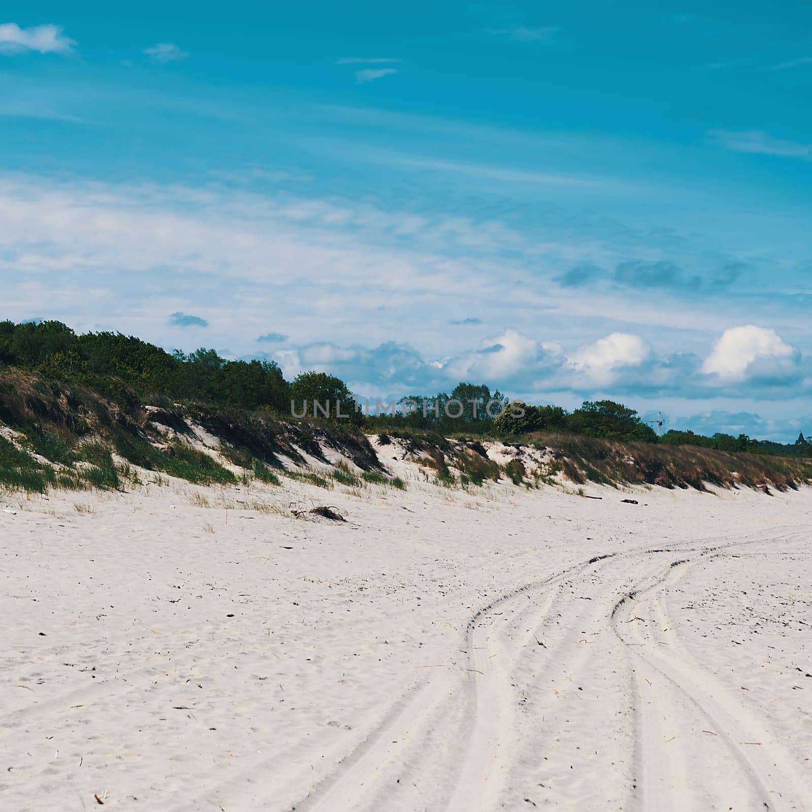 Dunes, overgrown with grass in places, car tracks on the sand. Blue sky with clouds.