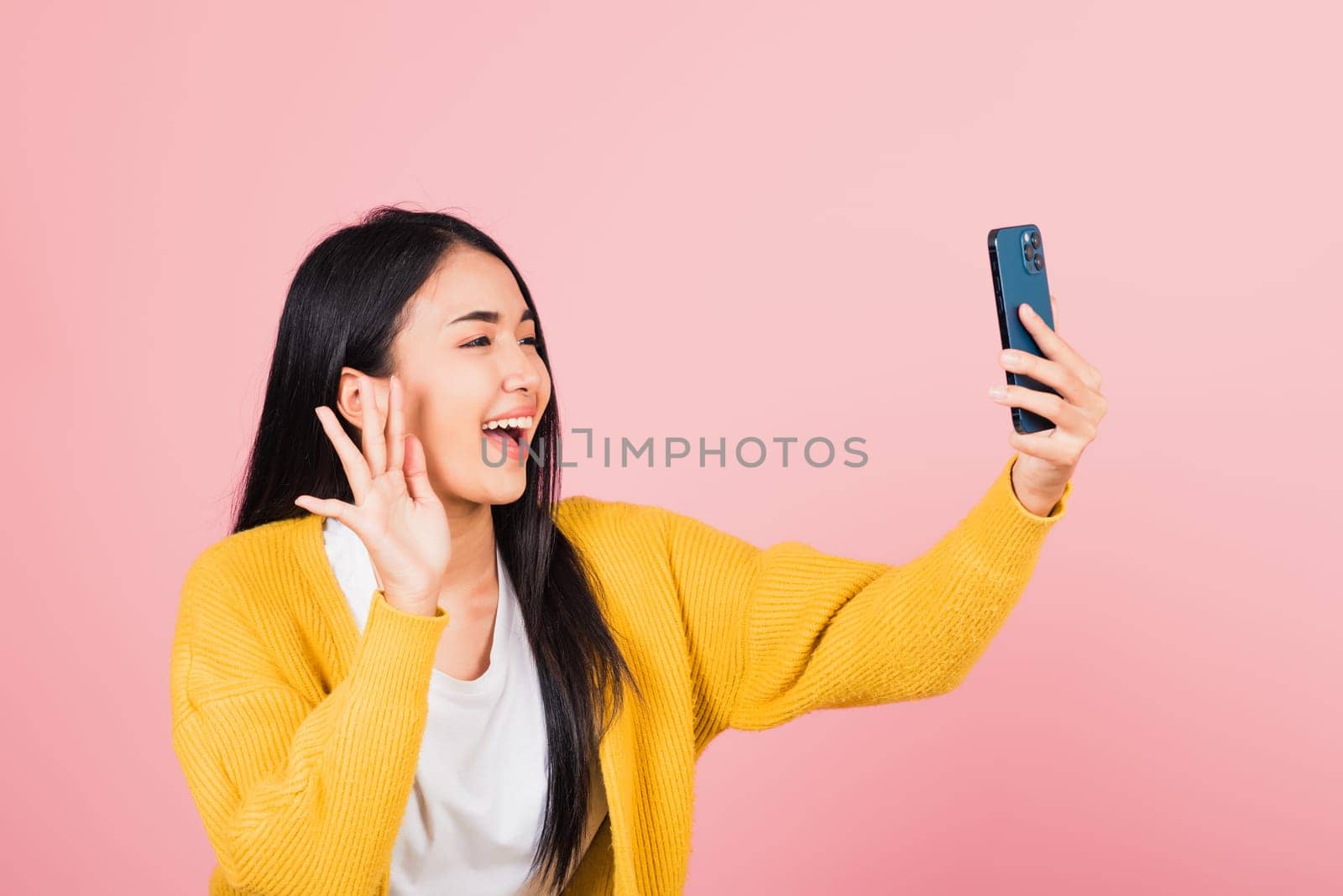 Happy Asian portrait beautiful cute young woman smiling excited  making selfie photo, video call on smartphone studio shot isolated on pink background, female hold mobile phone raise hand say hello