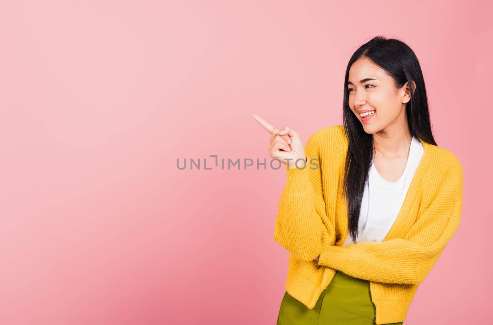 Asian happy portrait beautiful cute young woman standing pointing finger side away presenting product looking to away side, studio shot isolated on pink background with copy space