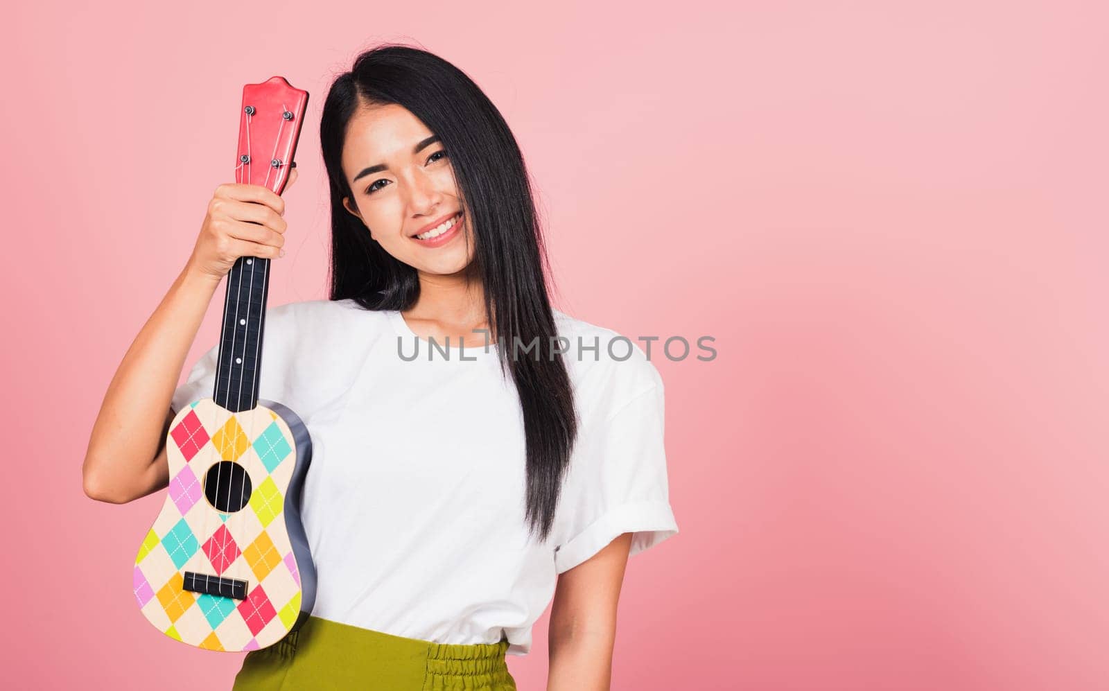 Portrait of happy Asian beautiful young woman teen confident smiling face hold acoustic Ukulele guitar, female playing Hawaiian small guitar, studio shot isolated on pink background, with copy space