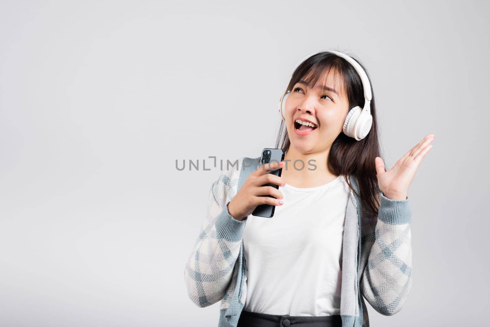 Woman excited smiling listening to music radio in bluetooth headphones and singing song on smartphone studio shot isolated white background, happy Asian young female Karaoke online app in mobile phone