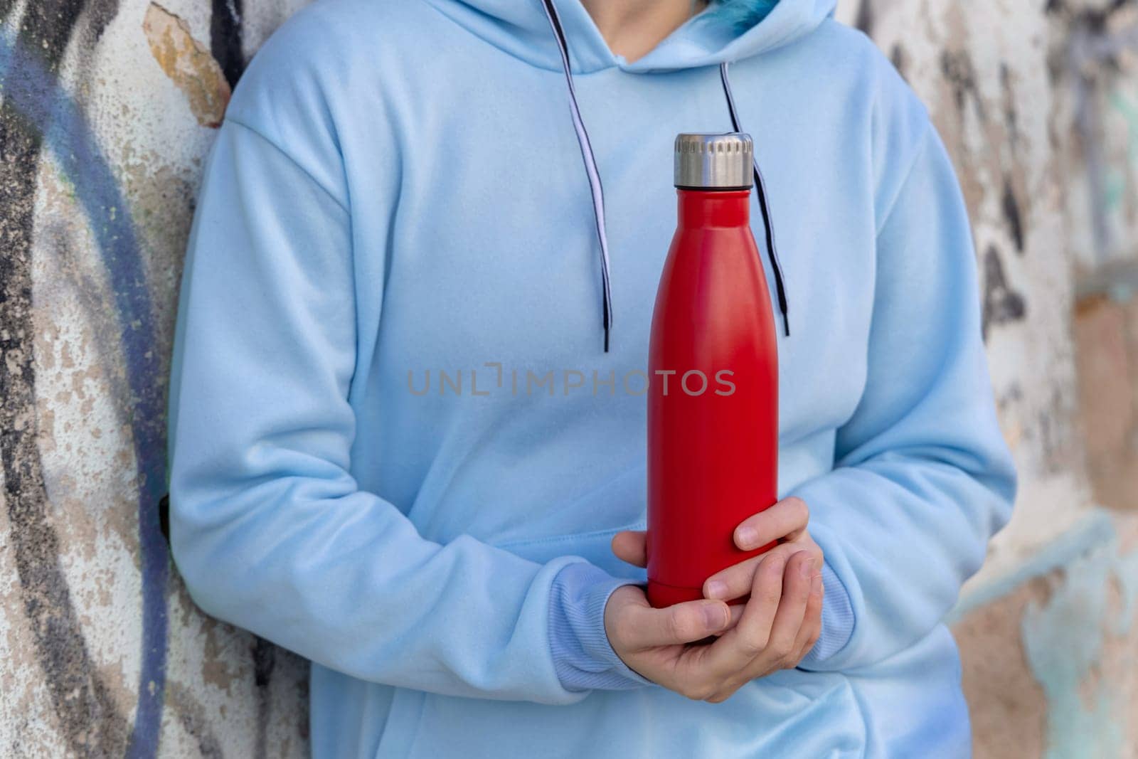 Young woman in blue hoodie with red water bottle staying outdoors near graffiti wall. Clothing and reusable bottle mockup. Hipster and adolescence concept
