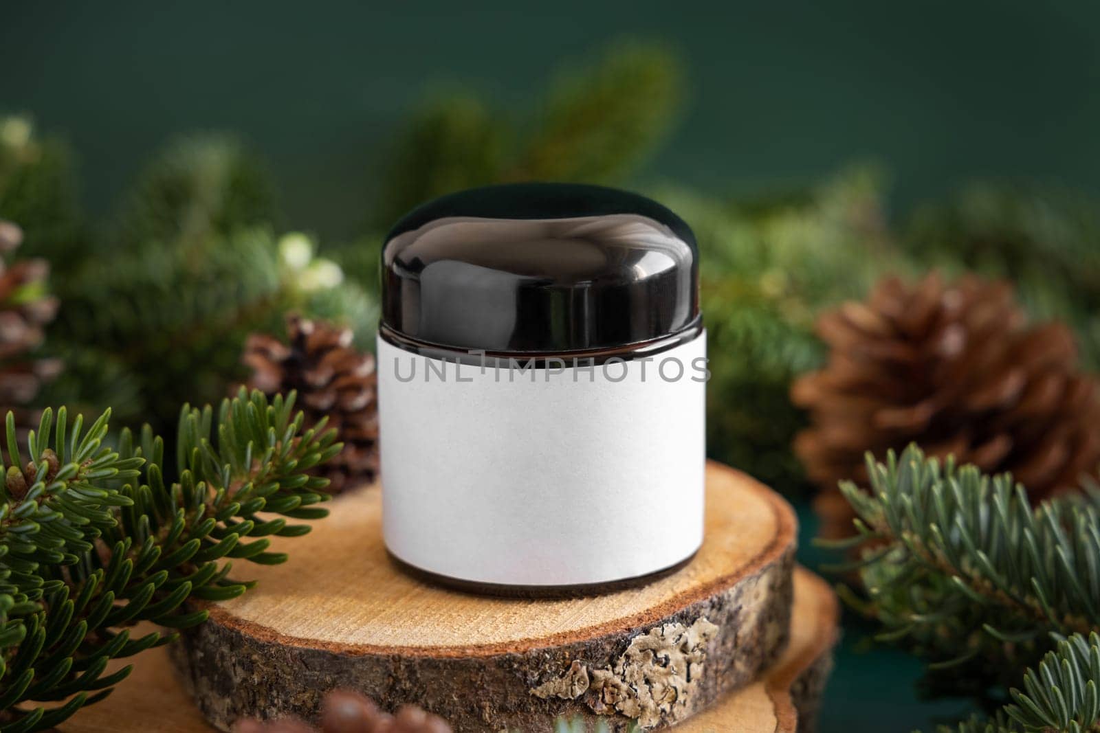 Glass jar between fir branches and pine cones on wooden piece close up. Brand packaging mockup. Dark cream container for skincare product, cream or serum. Healthcare concept. Natural cosmetics.