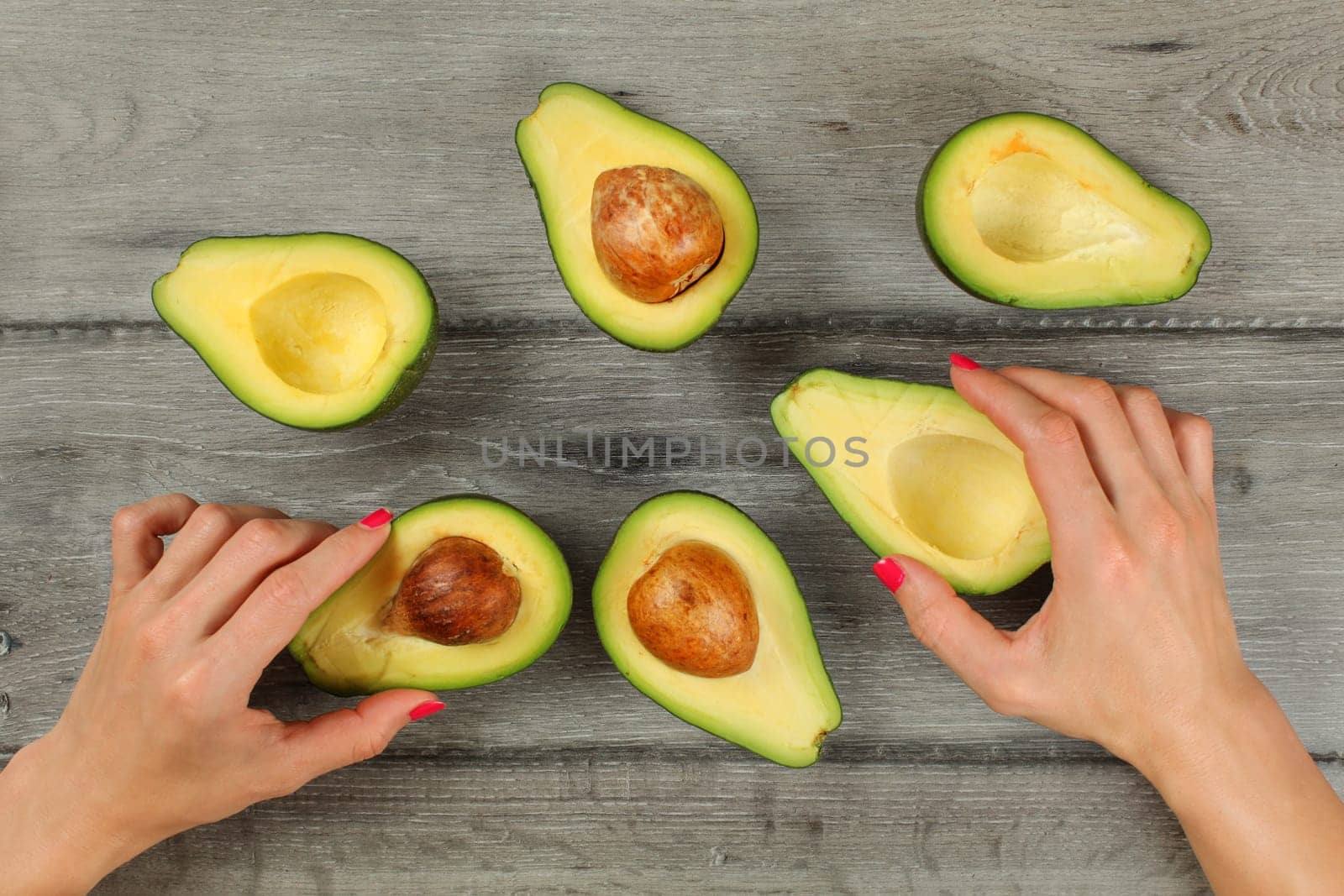 Tabletop view, woman hands with red nails holding avocados cut in half, more halves, some of them with seed around on gray wood desk.
