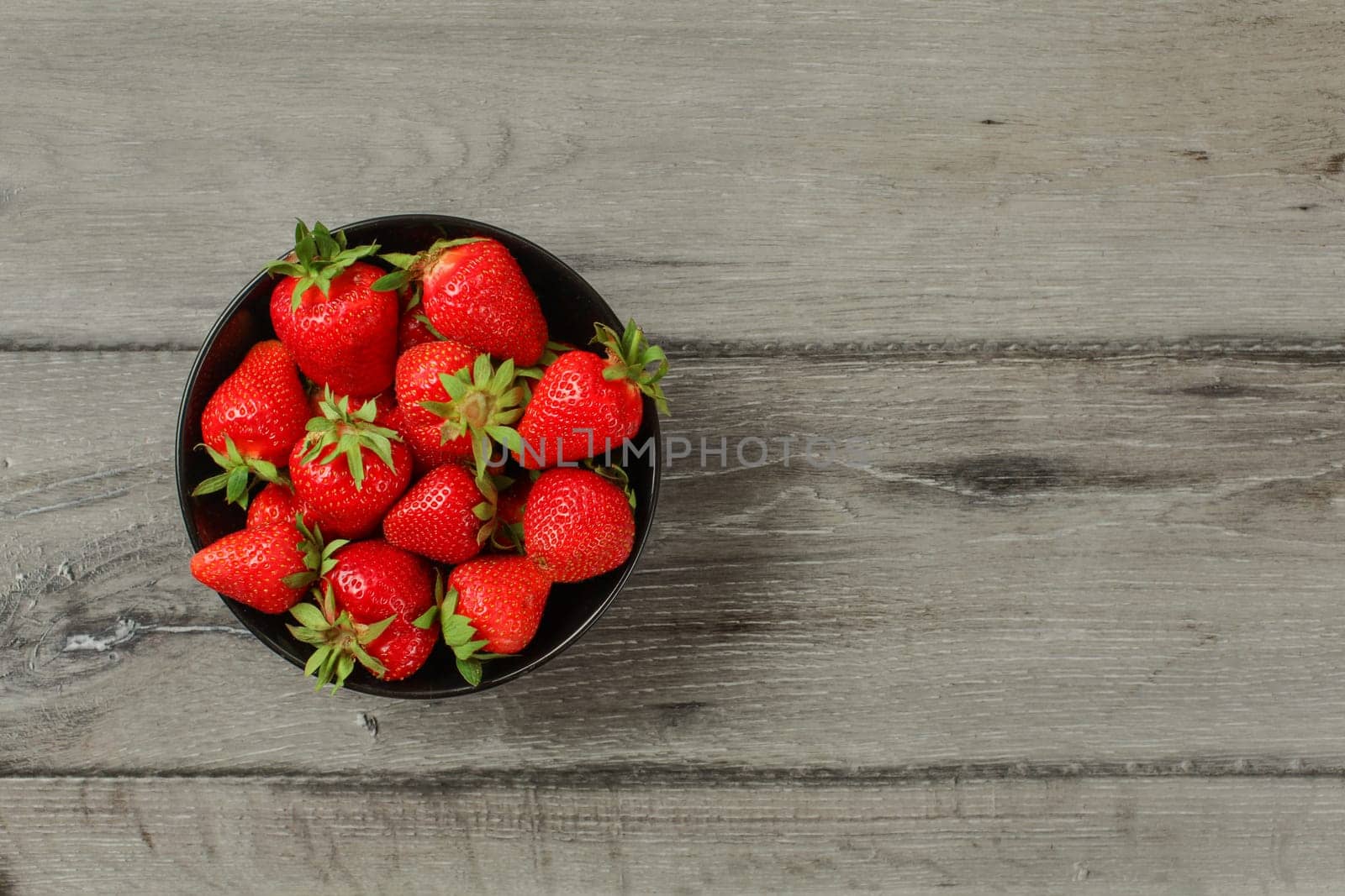 Tabletop view - small black ceramic bowl with freshly picked strawberries, on gray wood desk. Space for text on the right.