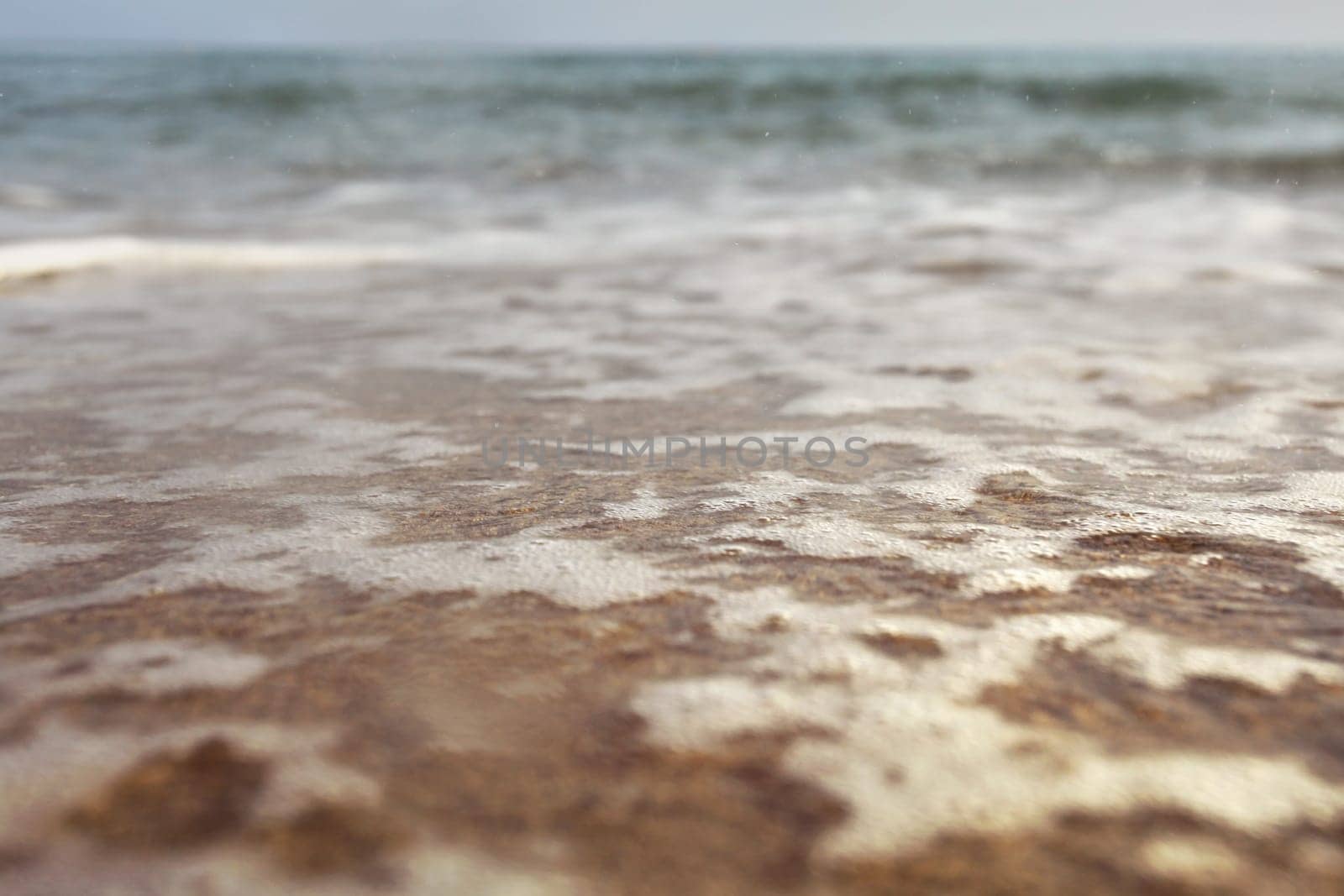 Low angle photo from ground level - beach with shallow water and white foam over sand, wet drops in air, blurred ocean in distance. Abstract vacation background.