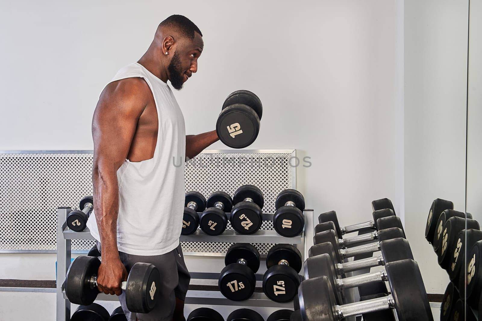 An African American man shakes his biceps with dumbbells while standing in front of a mirror. High quality photo