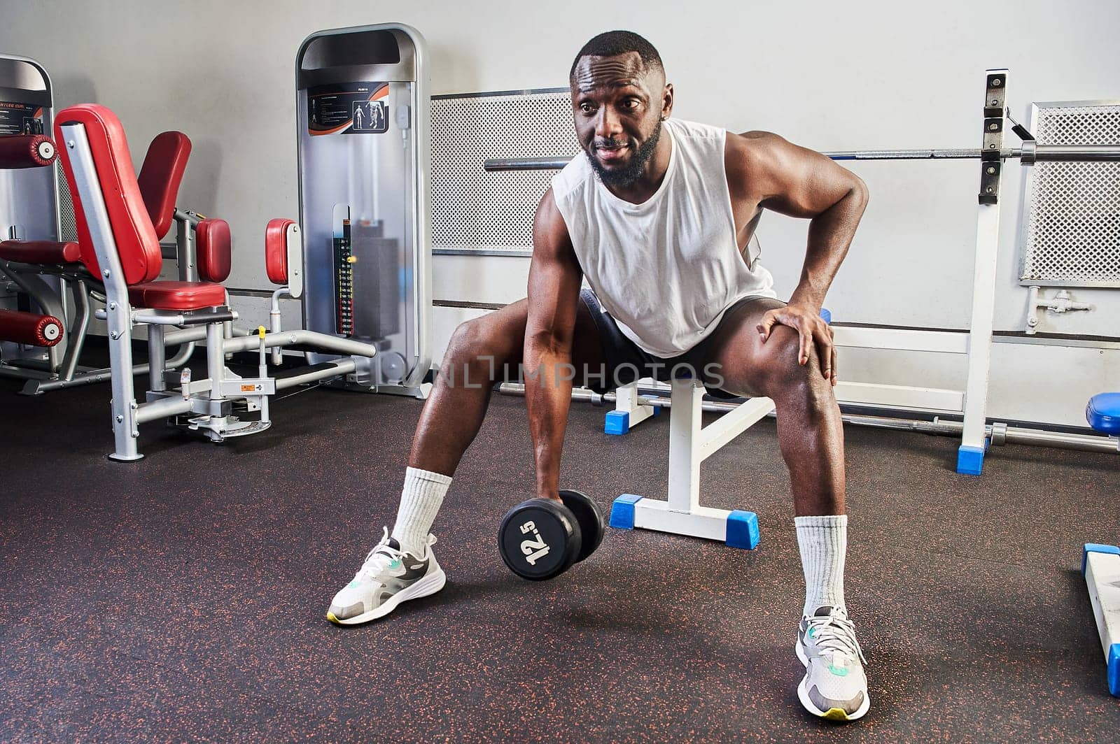 An African American man shakes his biceps while sitting on a bench in the gym. High quality photo