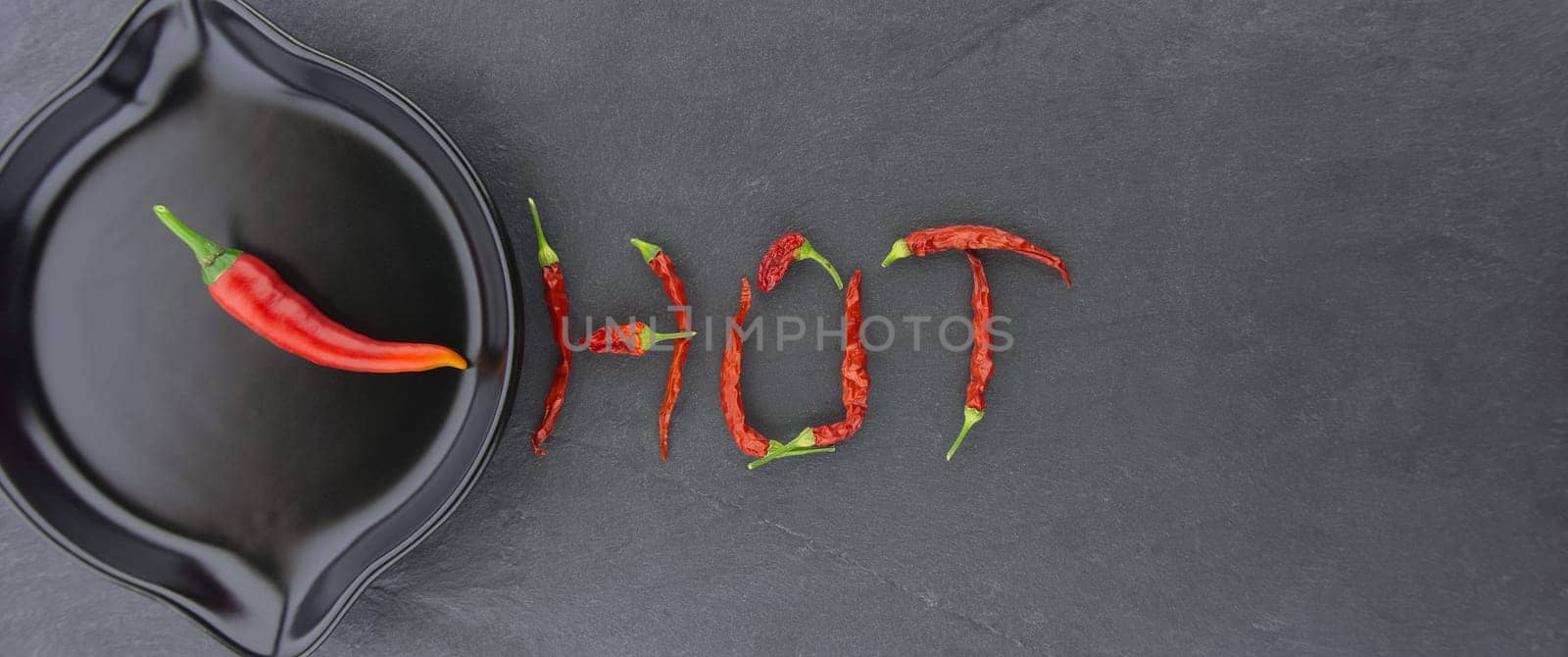 HOT spicy chili pepper still life with text formed from dried pods and a fresh chilli pepper in a skillet in a panorama banner over grey