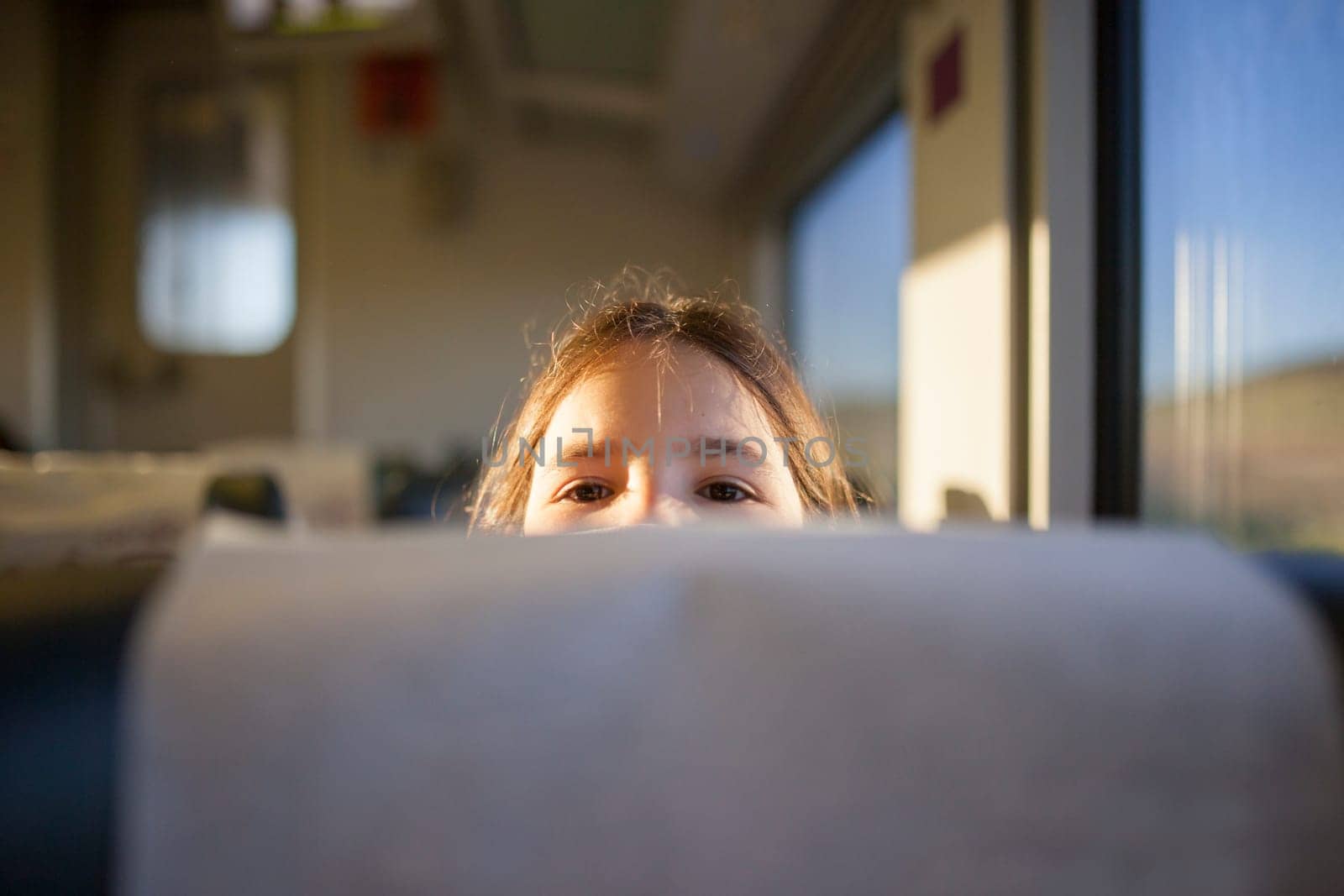 Child girl peeking out from behind a headrest train seat. Inside the train carriage