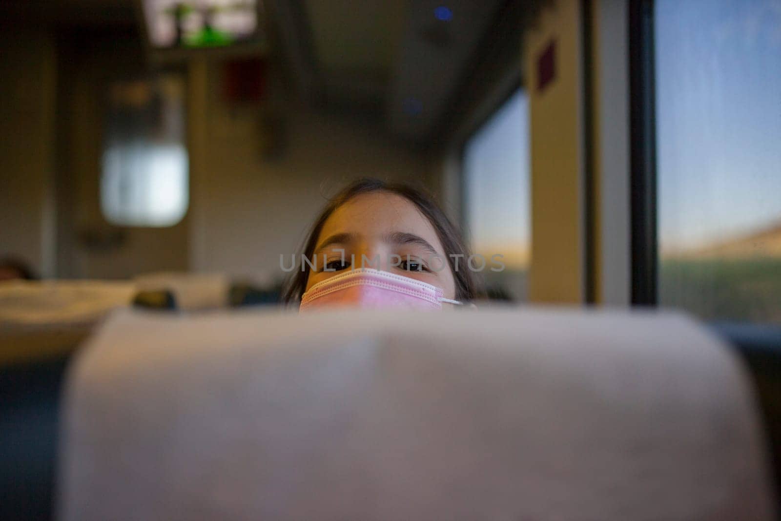 Child girl peeking out from behind a headrest train seat. Inside the train carriage