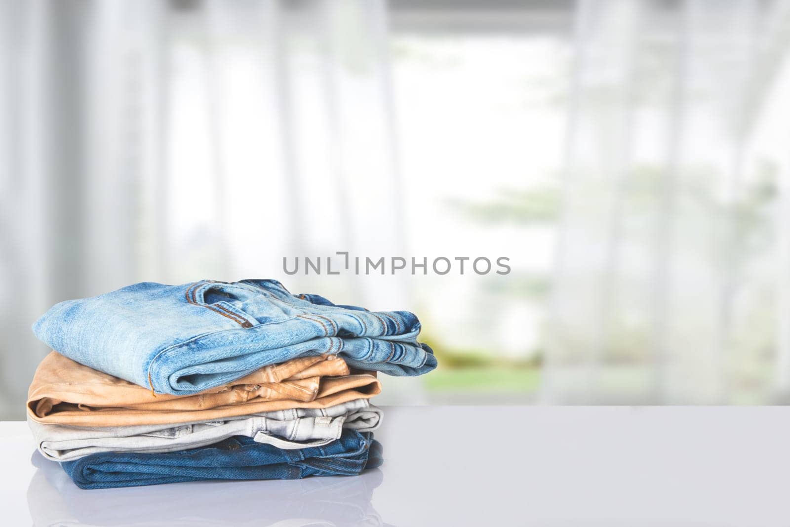 Stack of jeans. Closeup of a pile of colorful female denim pants on a bright table against abstract blurred curtain background. Copy space.