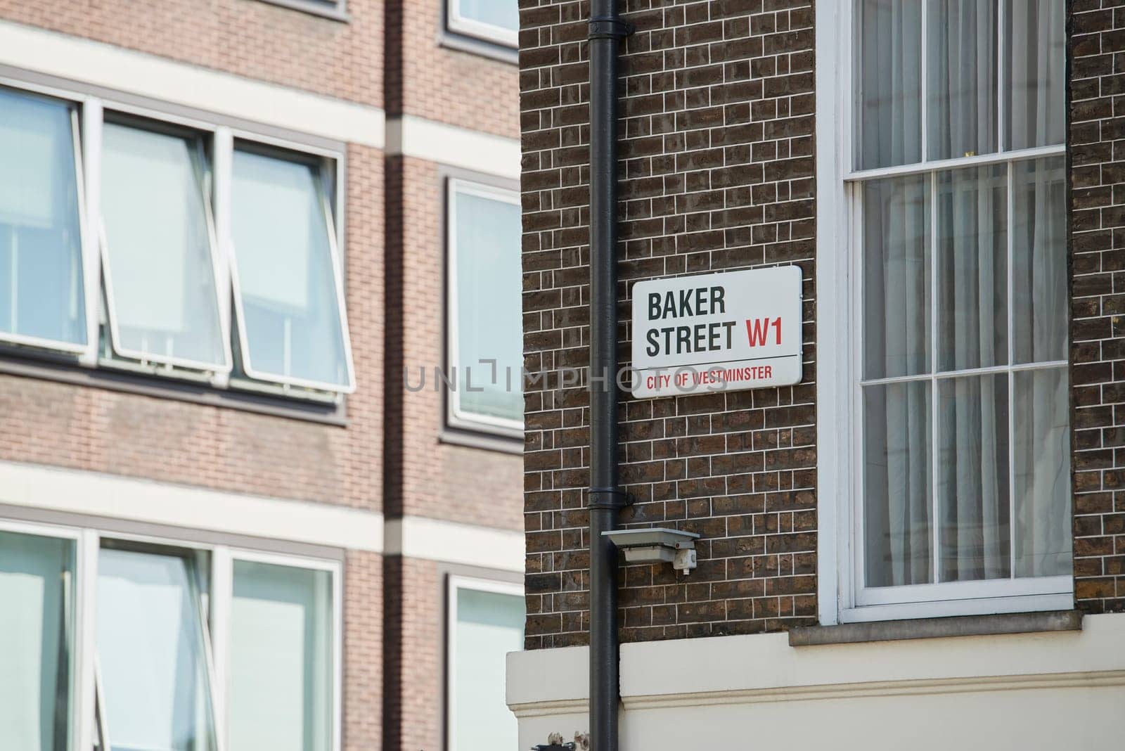 Baker Street sign affixed on dark brick wall next to drainage pipe and empty window. There are other building windows in the blurred background.