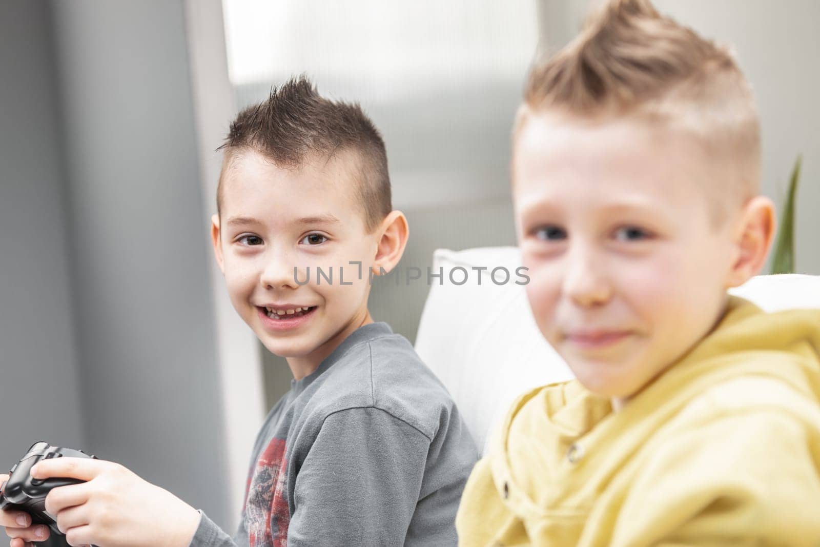 Cute mischievous brothers with spiky Mohican hairstyles sitting playing video games together on the sofa turning to smile at camera